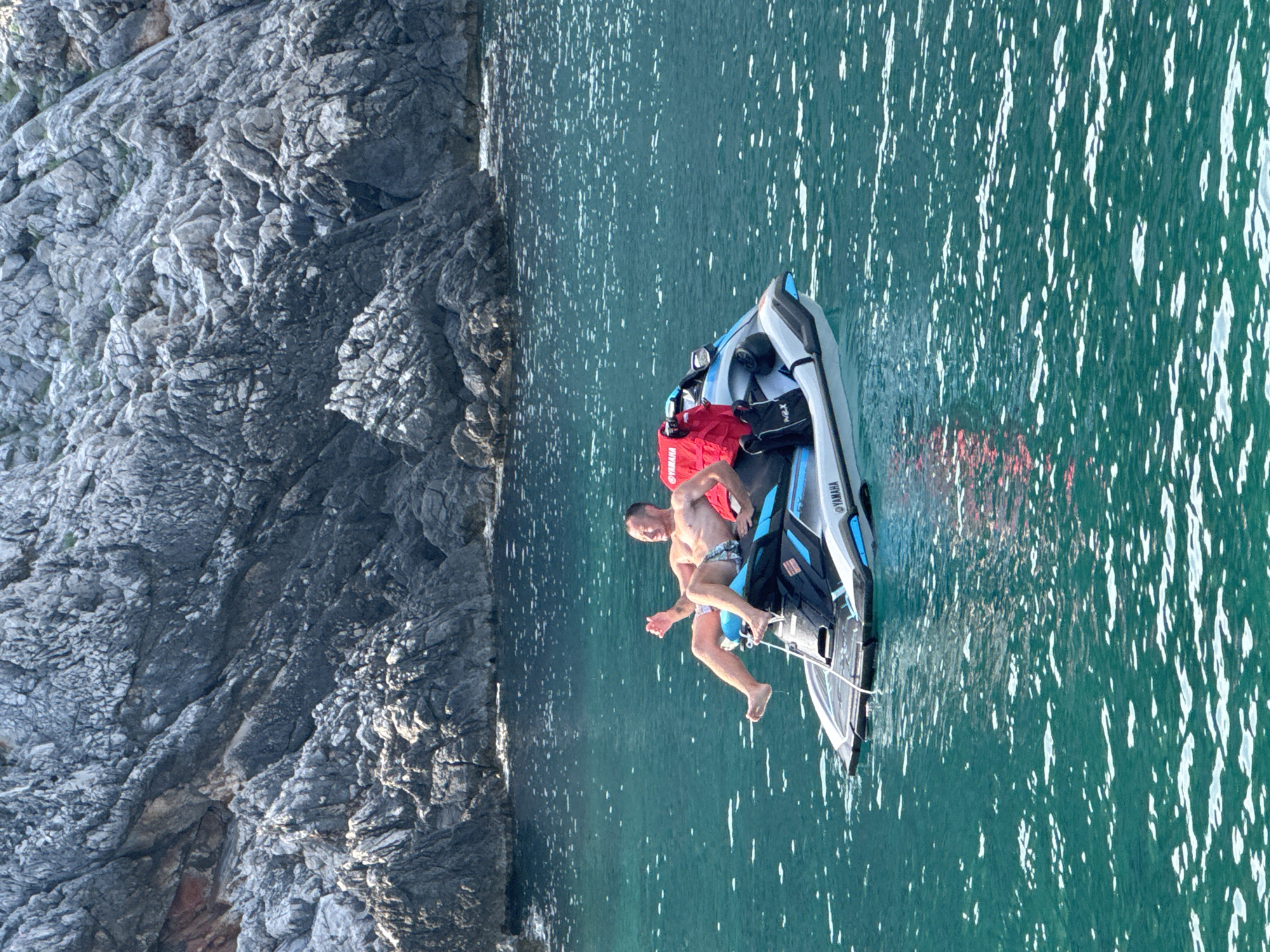 In Dubraljevina, Montenegro, a man is relaxing on a jet ski in clear turquoise water near a rocky coastline. The man, wearing checkered shorts, is leaning back on the jet ski with his legs extended and his left arm raised. A red bag is secured behind him on the jet ski. The water is slightly rippled, with sunlight glinting off the surface. The rocky shore, characterized by its grey, textured surface, forms the backdrop to the scene. The jet ski has "YAMAHA" branding visible on its side.