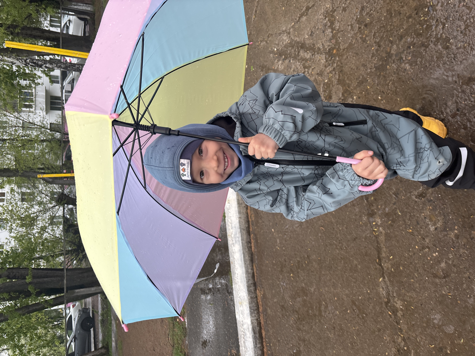 A young child stands outside under a colorful umbrella. The child is wearing a gray hooded jacket and yellow boots, and holds the pink handle of the umbrella. The background includes buildings and trees, a curb, and a wet asphalt surface. The location is Chaikovsky, Russia.