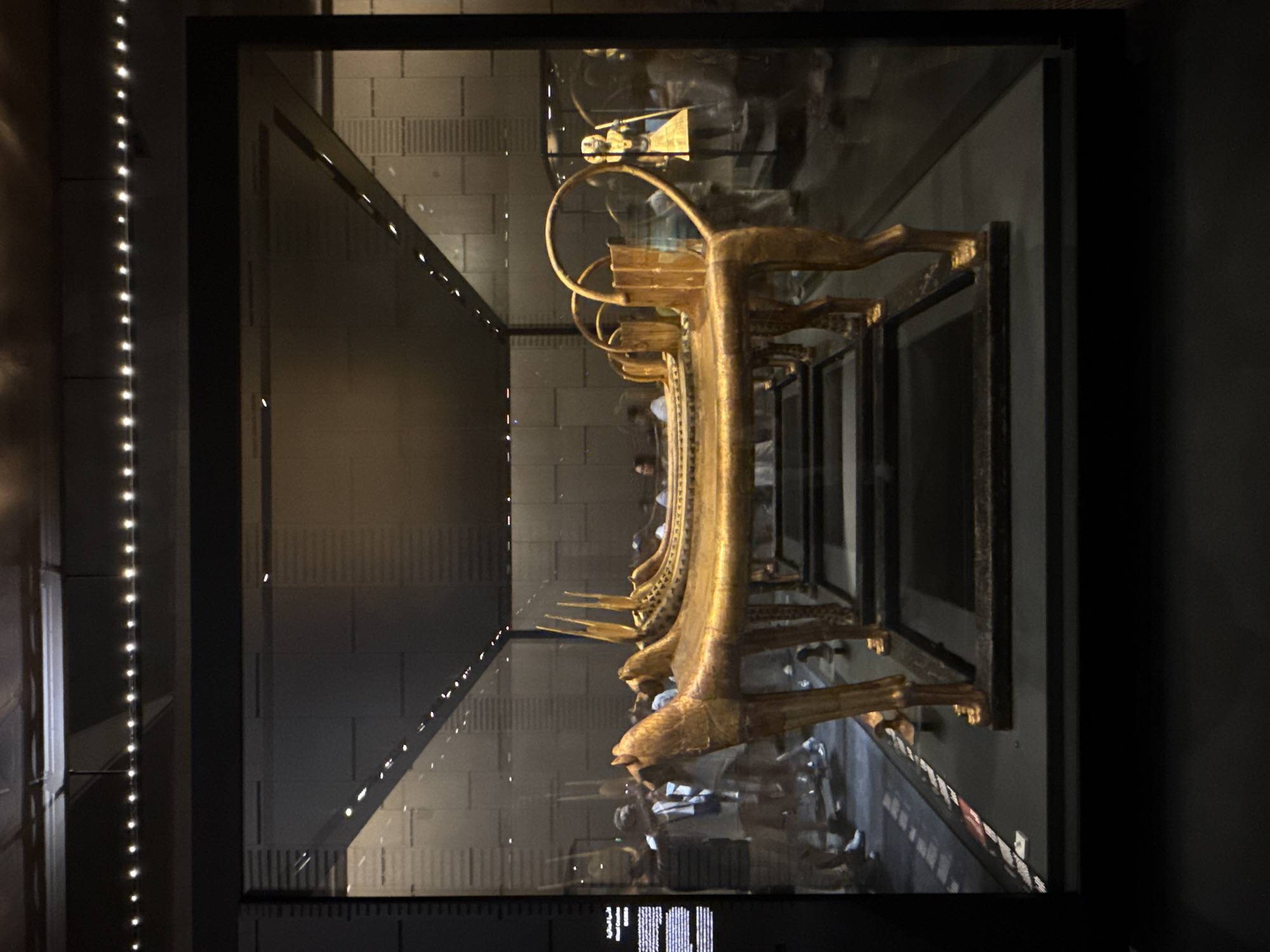 The image displays a display case within a museum setting, likely housing ancient artifacts. Prominently featured are several ornate, golden ancient Egyptian funerary couches, each supported by legs shaped like animals, possibly lions or horses. These couches appear to be arranged in a row within the glass enclosure. In the background, through the glass, several people are visible, observing the exhibits. Further in the distance, another display case holds a golden statue of a figure in a characteristic Egyptian headdress and attire. The lighting in the museum is subdued, with a string of small, white lights visible along the top edge of the display case and reflected on the ceiling. The background walls are dark and feature a textured, possibly gridded, pattern. The floor of the display area is dark, with a red and white striped border visible near the front. The overall impression is of a well-lit, curated museum exhibition of significant historical importance. The location provided, Kafrat Nassar, Egypt, is a village in the Giza Governorate, which is home to the Giza pyramid complex and numerous other ancient Egyptian sites, making it a plausible location for such a museum.