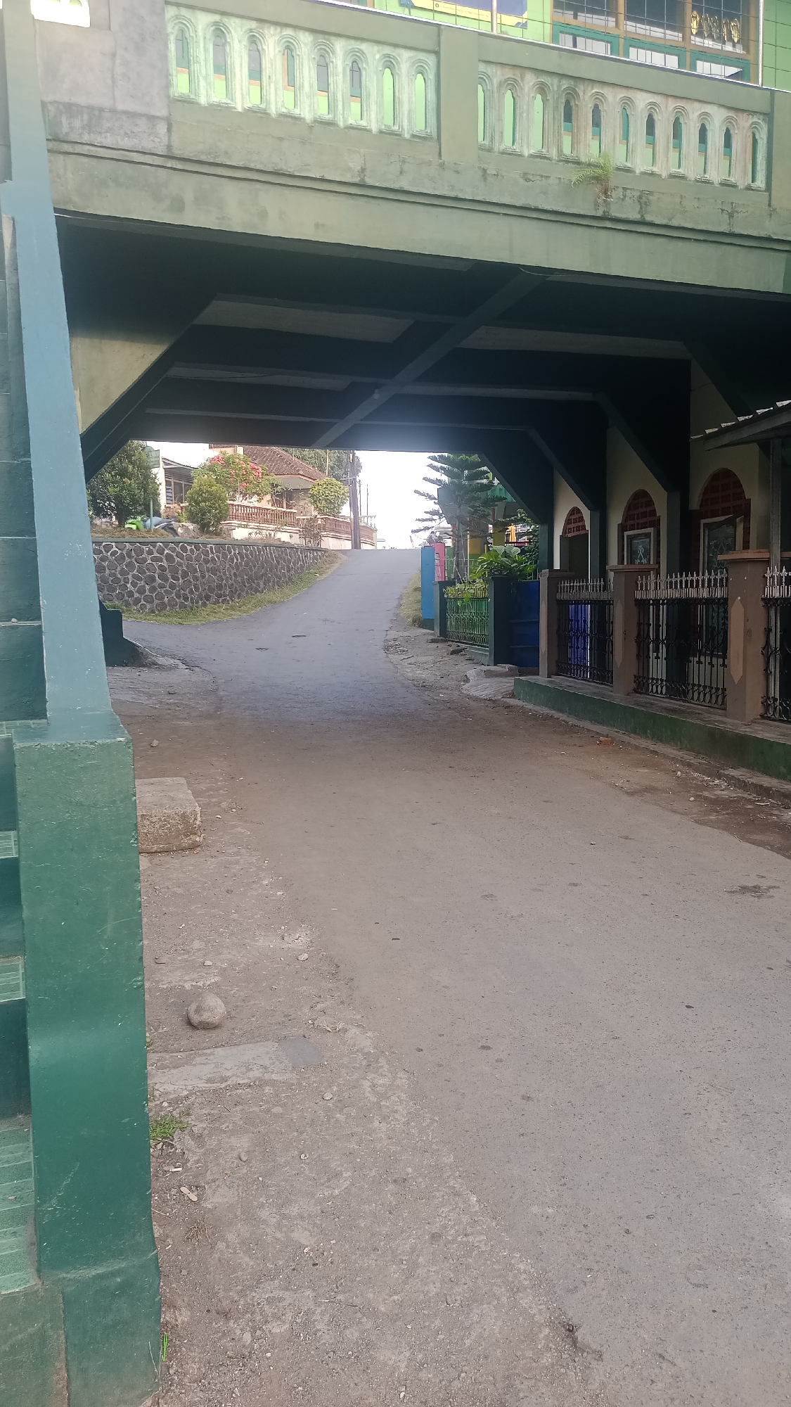 This image depicts a view of a street passing under a green bridge or overpass, likely in Karyamandala, Indonesia. The bridge structure is prominent in the foreground and upper portion of the image, with decorative green and white railings visible on its upper side. Beneath the bridge, the street curves gently upwards and to the left. On the left side of the street, a low stone wall borders a raised area with vegetation and a house. On the right side of the street, there is a building with a patterned facade, featuring arches and ornamental fencing. Various trees and fences are also visible along the sides of the street, adding to the residential or urban landscape. The overall impression is of a quiet, possibly suburban street in a tropical or subtropical region.