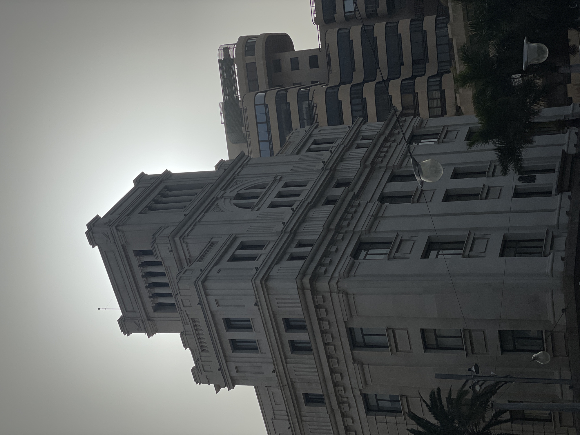 The image shows a low-angle shot of two buildings under a bright, hazy sky. The primary building, in the foreground, has a classical architectural style with multiple stories, each defined by horizontal lines of windows and decorative molding. The building is topped with a tower featuring columns and a rounded window detail. A smaller antenna extends from the top of the tower. Behind this building, a taller, more modern structure is visible. Its design includes curved, protruding sections with dark-tinted windows. Palm trees and streetlights are visible on the right side of the frame. The scene appears to be set in the city of Santa Cruz de Tenerife, Spain.