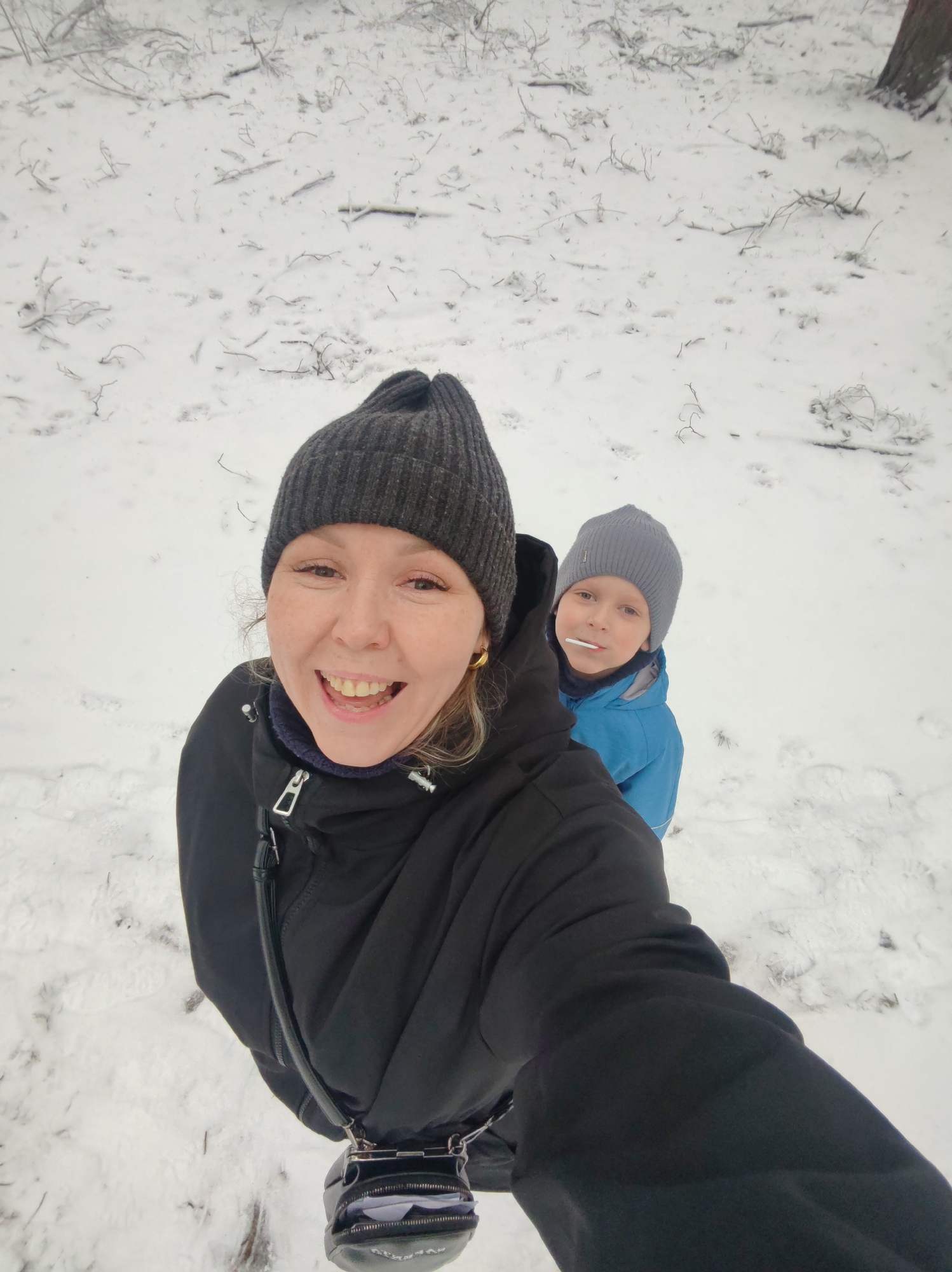In a winter scene in Chaikovsky, Russia, a smiling woman in a black jacket and grey knitted hat takes a selfie. Standing behind her is a young boy in a grey knitted hat and blue jacket, holding a lollipop in his mouth. The ground is covered in snow, with sparse vegetation visible beneath.
