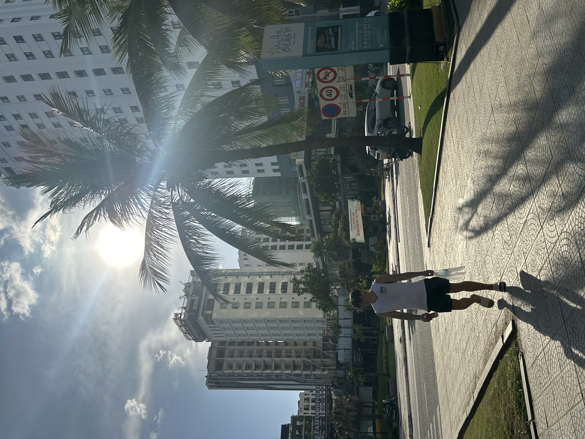 The image captures a street scene in the city of Đà Nẵng, Vietnam, on a sunny day with a bright sun and partial cloud cover. In the foreground, a young man is walking on a paved sidewalk, casting a long shadow. He is wearing a white sleeveless shirt and dark shorts, with flip-flops on his feet. He is carrying a red cylindrical object in his left hand and a white plastic bag in his right.
Behind him, a road runs parallel to the sidewalk, with a silver car visible on the right side of the road. Further back, there are several tall buildings, some appearing to be under construction, with modern architecture. Palm trees are scattered throughout the scene, with their fronds partially obscuring the view of the buildings and sky. A blue and white sign with Vietnamese text is visible on the right side of the image, appearing to be related to a beach or tourist destination. Traffic signs, including one indicating a speed limit of 40, are also present. The overall impression is a vibrant urban environment with tropical elements.