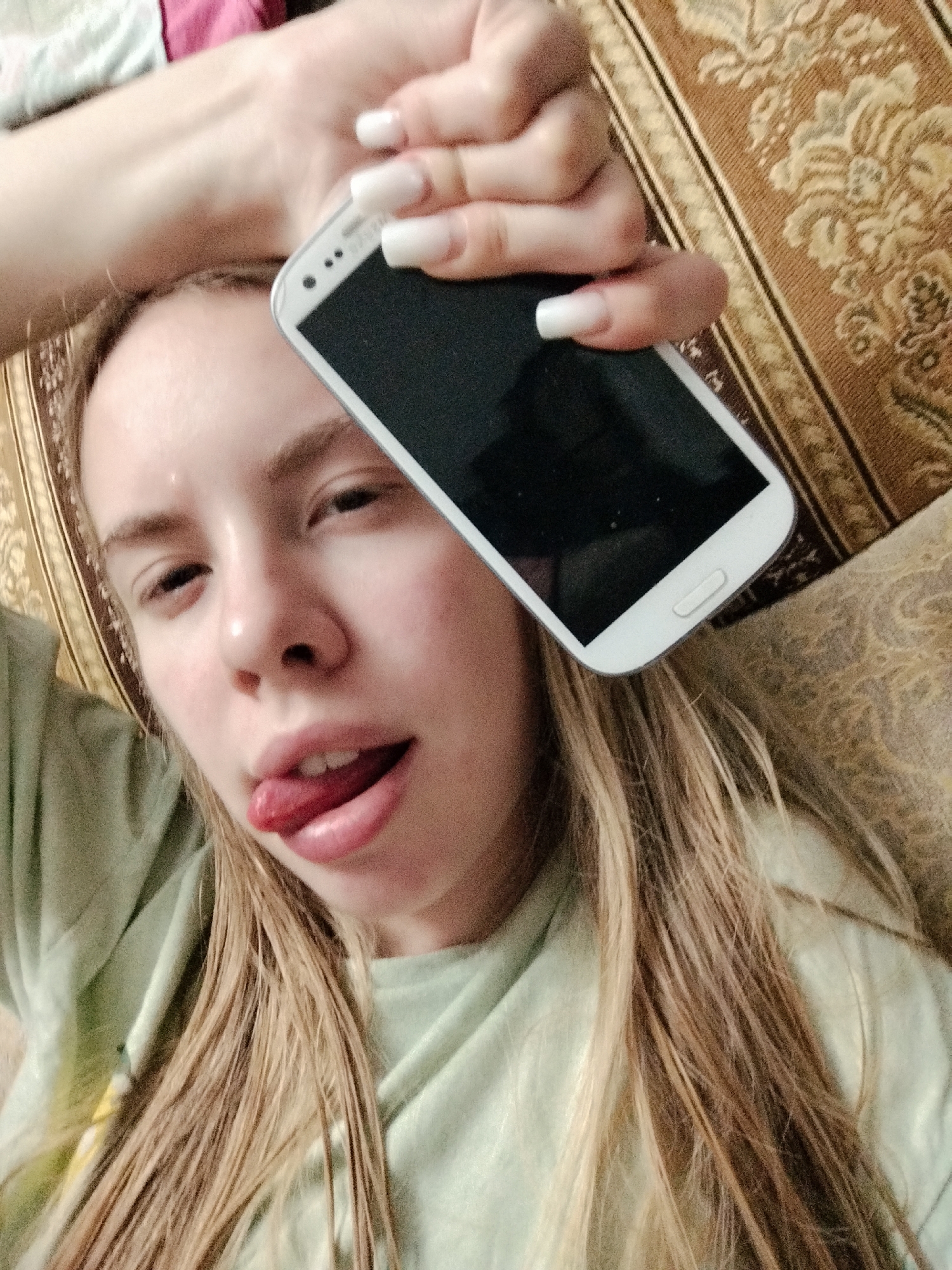 An overhead, close-up, indoor, selfie-style photograph shows a young Caucasian female with blonde hair lying on her back on a patterned couch and holding a white smartphone over her face. The female's left arm is bent and raised above her head, with her hand holding the phone to obscure the top portion of her face. She is wearing a light green t-shirt. Her tongue is sticking out of her mouth, and her eyes are partially closed. The background consists of the textured fabric of the couch. The lighting appears to be artificial.