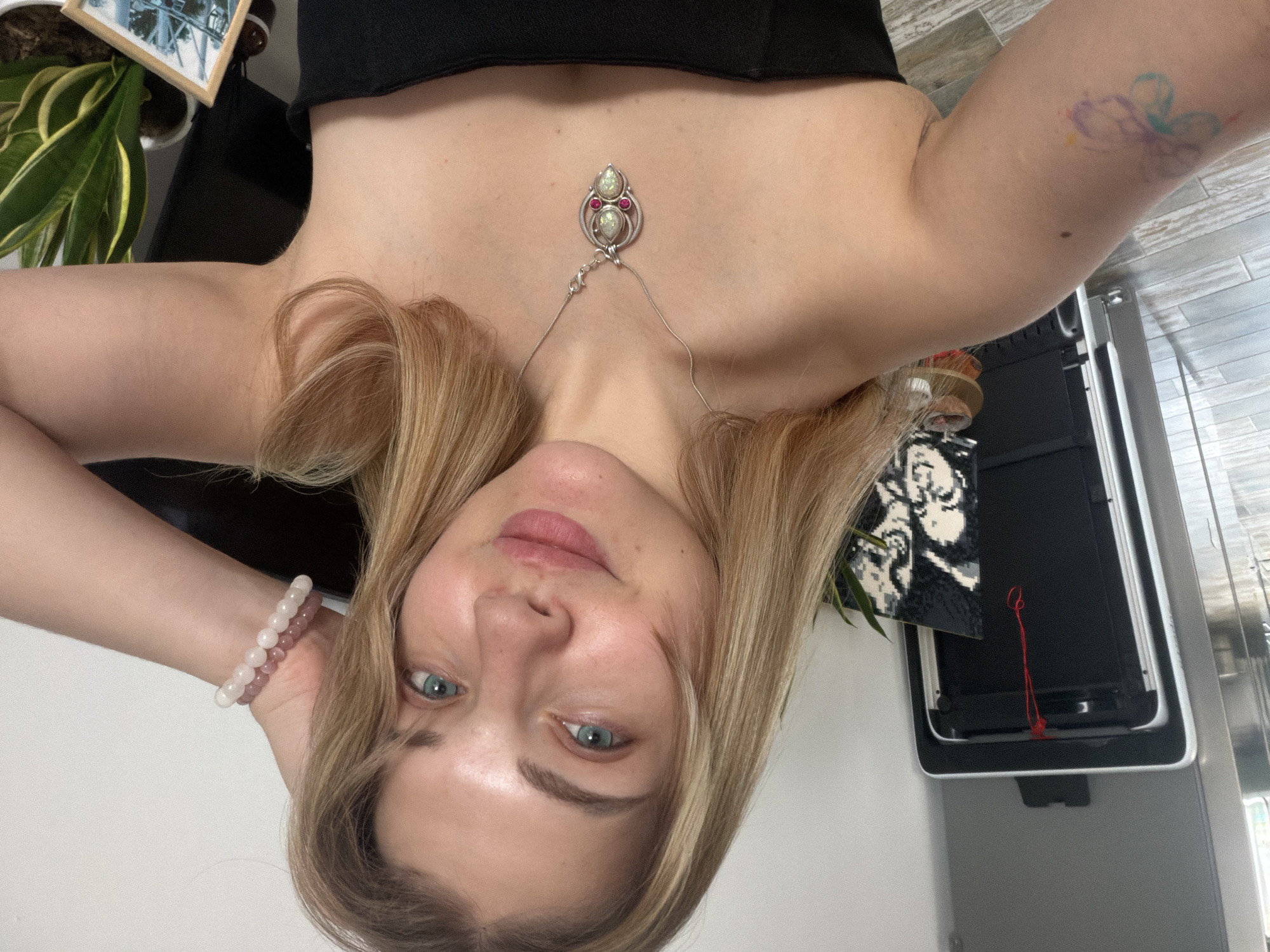 A woman is taking a selfie. The image appears to be taken indoors. She is wearing a black top and a necklace with a decorative pendant. She is wearing beaded bracelets on her left wrist. A plant and a framed picture are visible in the background. A white wall and part of what appears to be a kitchen appliance can be seen. There is no information to indicate that the photograph was taken in Большая Шильна, Russia.