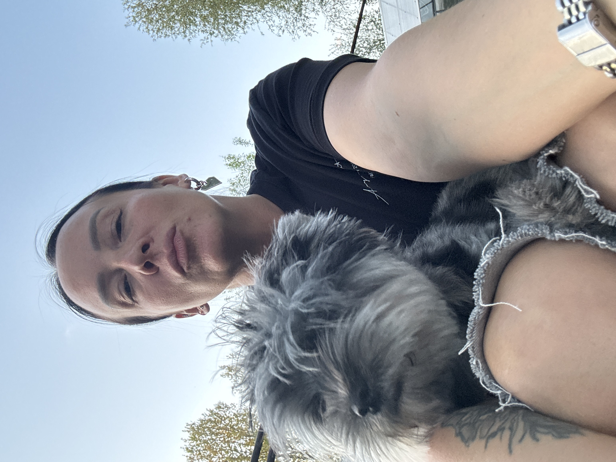A person, likely female, is holding a small, gray, fluffy dog. The person is wearing a black t-shirt and has a silver wristwatch on their left wrist. They also have earrings. The dog is resting in their lap, which is covered with what appears to be denim clothing with frayed edges. The background includes a view of the sky and trees. The location is not identifiable as Chaikovsky, Russia, based on the image.