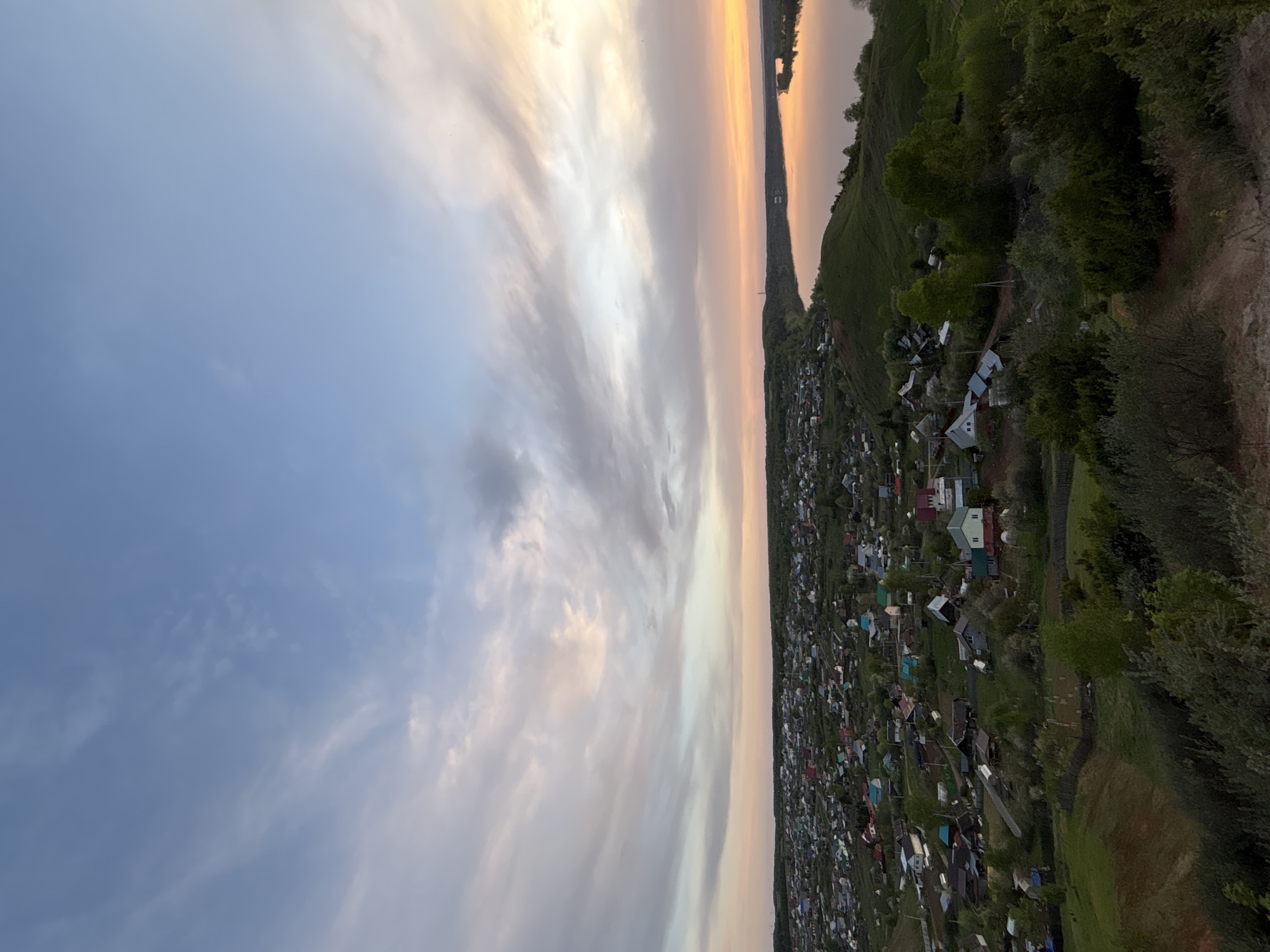 The image shows a landscape in Ольховка, Russia, likely taken at sunset. A body of water, possibly a river or lake, is visible in the distance, with its surface reflecting the colors of the sky. To the right of the water is a green, tree-covered hill, leading down to a densely populated area with numerous houses and buildings. The sky occupies a large portion of the image, displaying a mix of blue and pink hues, indicative of a sunset or sunrise. Clouds are scattered across the sky, adding texture and depth to the scene.