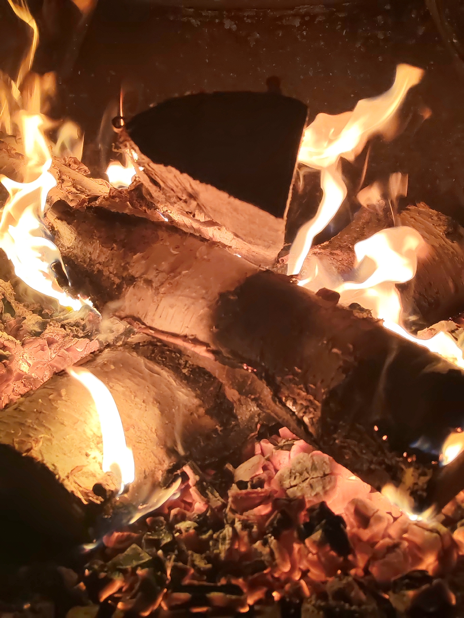 The image depicts a close-up view of a fire, presumably in a fireplace or fire pit. Several logs are visibly burning, with flames extending upwards and around them. Glowing embers are present at the bottom of the fire. The location is Unknown, Unknown.