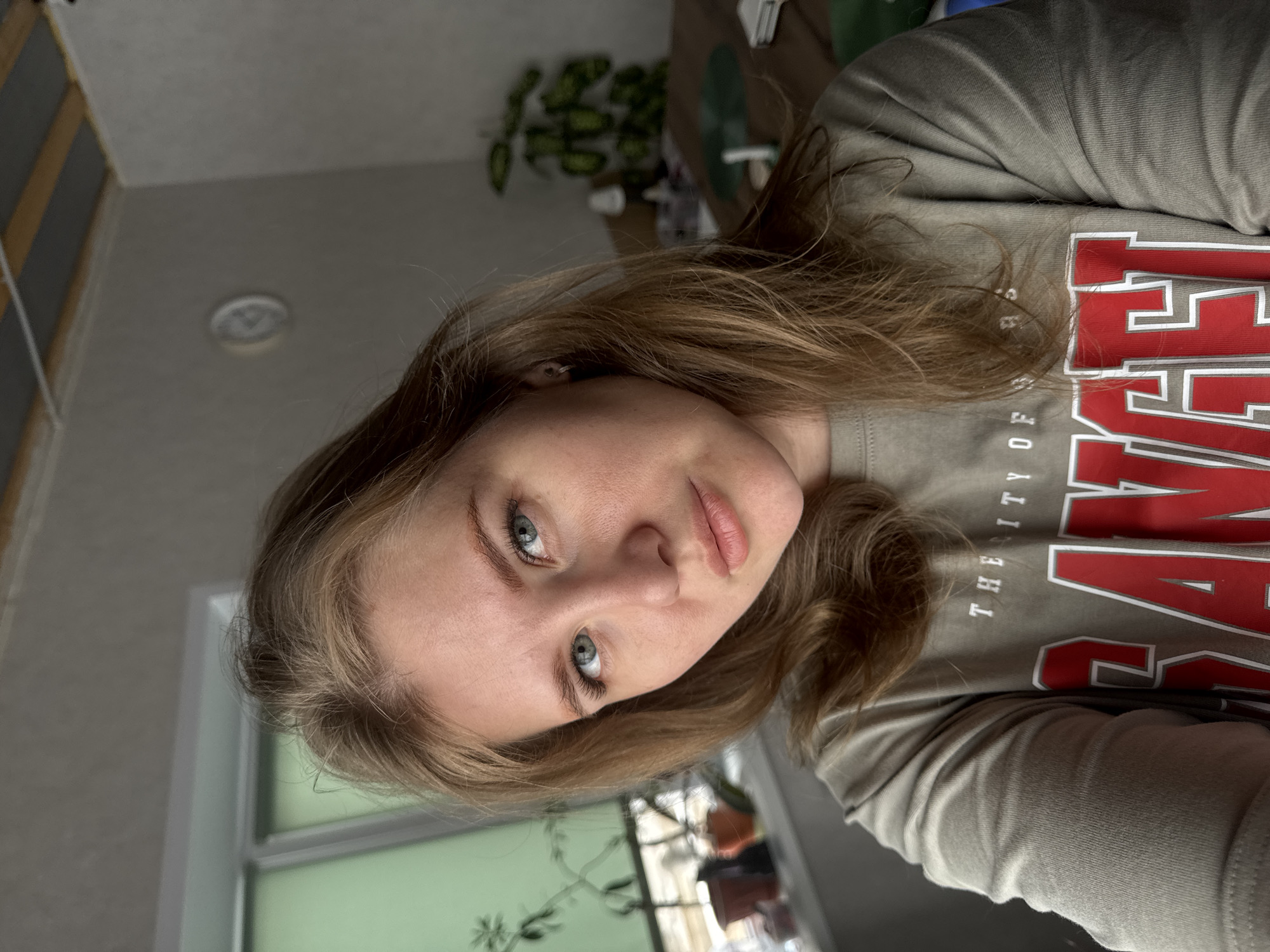 A close-up, angled shot shows a fair-skinned young woman with light brown hair and blue eyes. She wears a grey long-sleeved shirt with the word "THE CITY OF" and a large red letter visible. The background includes a window with a visible plant and a wall with a ceiling light fixture. The location in Gornyi (Dvurechenskoe S/P), Russia is not directly discernible from the image content.