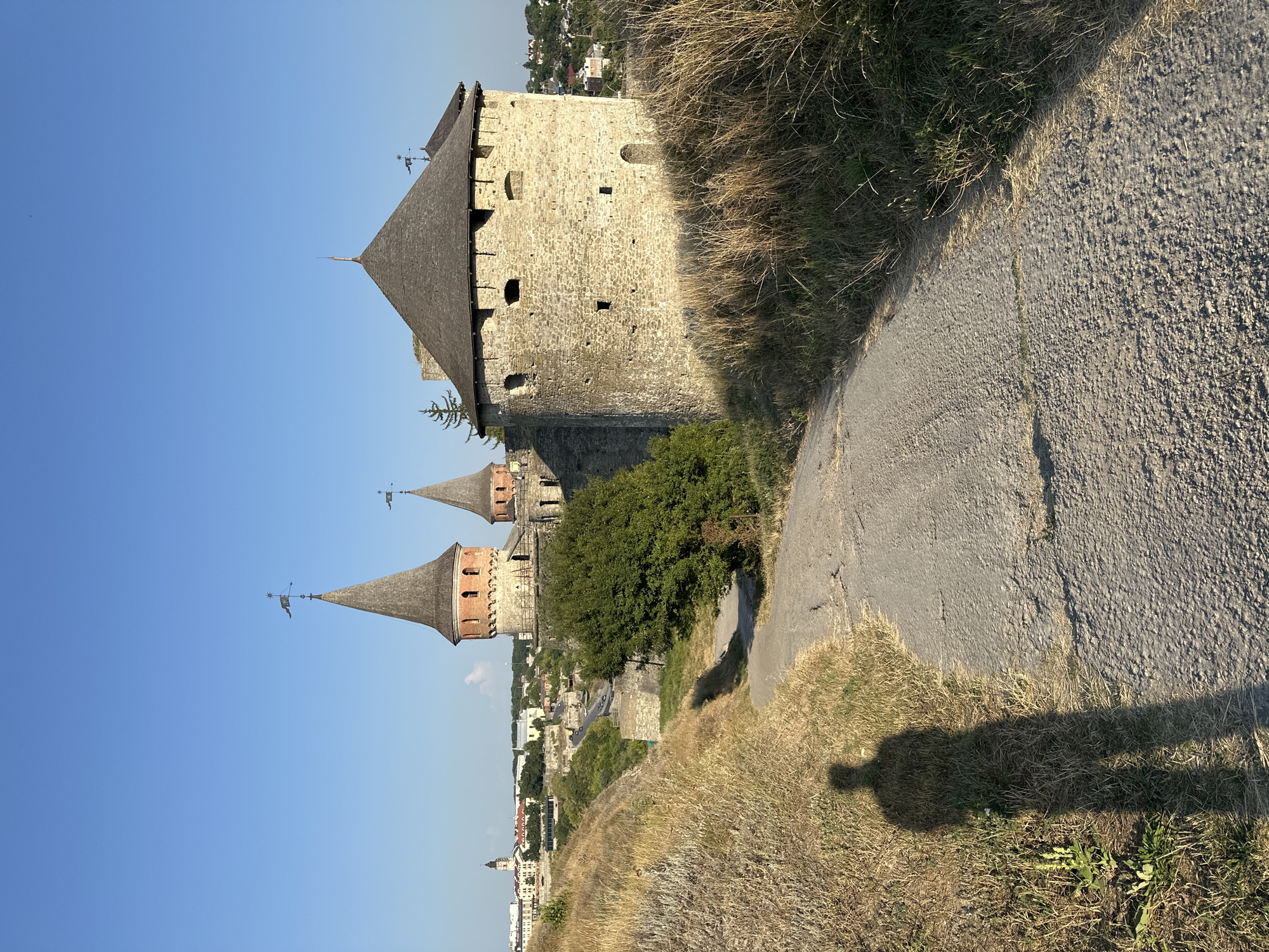 The image captures a view of the Kamianets-Podilskyi Castle in Ukraine, a prominent historical landmark situated on a rocky promontory. In the foreground, a paved path winds its way up a grassy incline. The shadow of a person is cast on the left side of the path. The castle itself is visible in the middle ground, characterized by its stone walls and conical-roofed towers. One tower has a weather vane topped with a flag. Beyond the castle, the cityscape of Kamianets-Podilskyi unfolds, with buildings and church spires under a clear blue sky. The surrounding landscape is dotted with trees and dry grass, indicative of a sunny day.