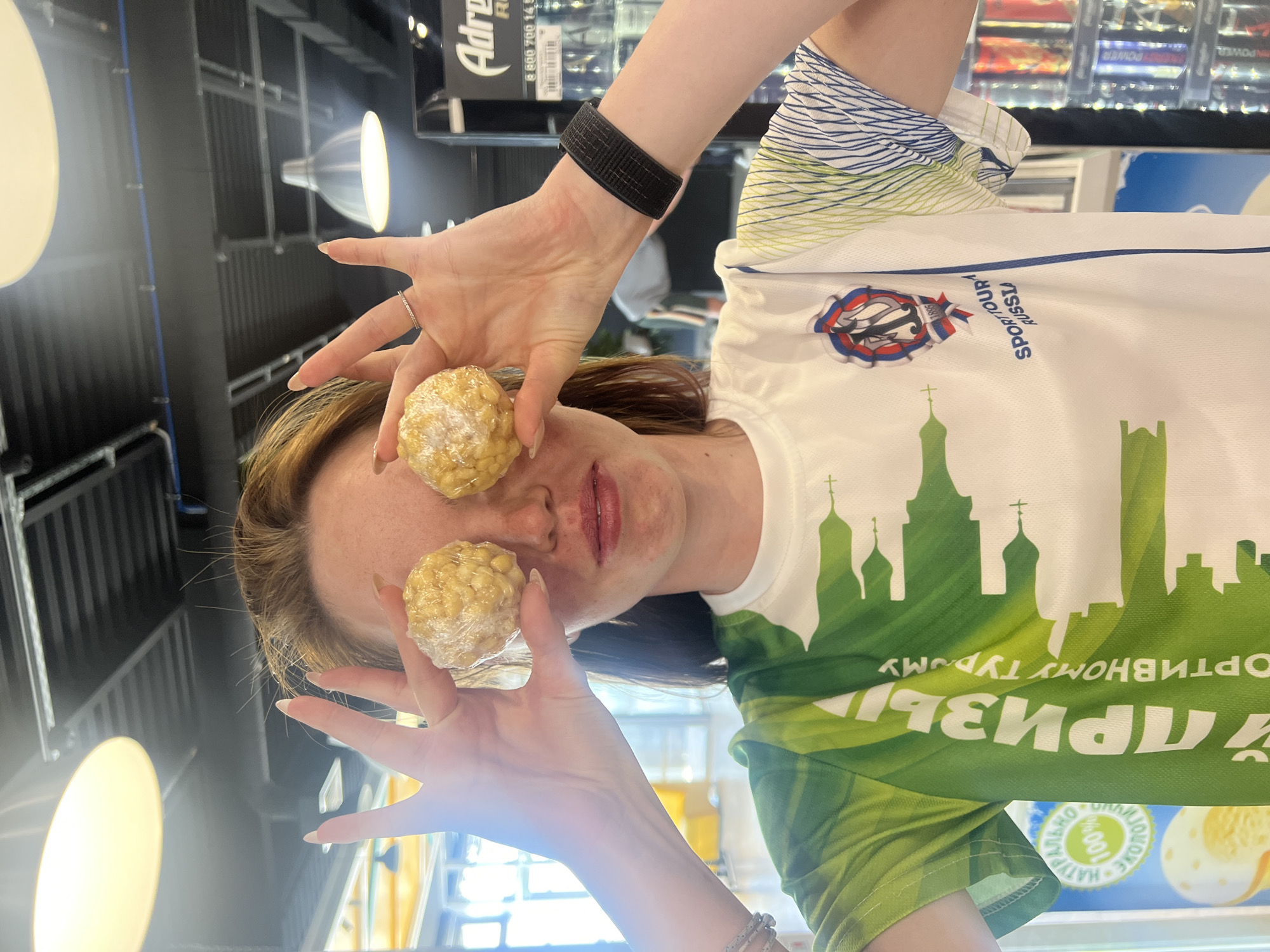 The image shows a woman holding two wrapped rice crispy treat balls in front of her eyes, as if they are her eyes. She is indoors, likely in a shop or store. She is wearing a white t-shirt with green and blue accents, featuring an emblem with the text "SPORT TOUR RUSSU" and a design that resembles a cityscape with churches. The shirt also has Cyrillic text that appears to be related to travel or prizes. Her left wrist has a black activity tracker, and there's a ring on one of her fingers. The background has overhead lighting and a display of products, possibly snacks or drinks. Based on the church silhouette on the shirt, it is potentially in Казань, Russia.
