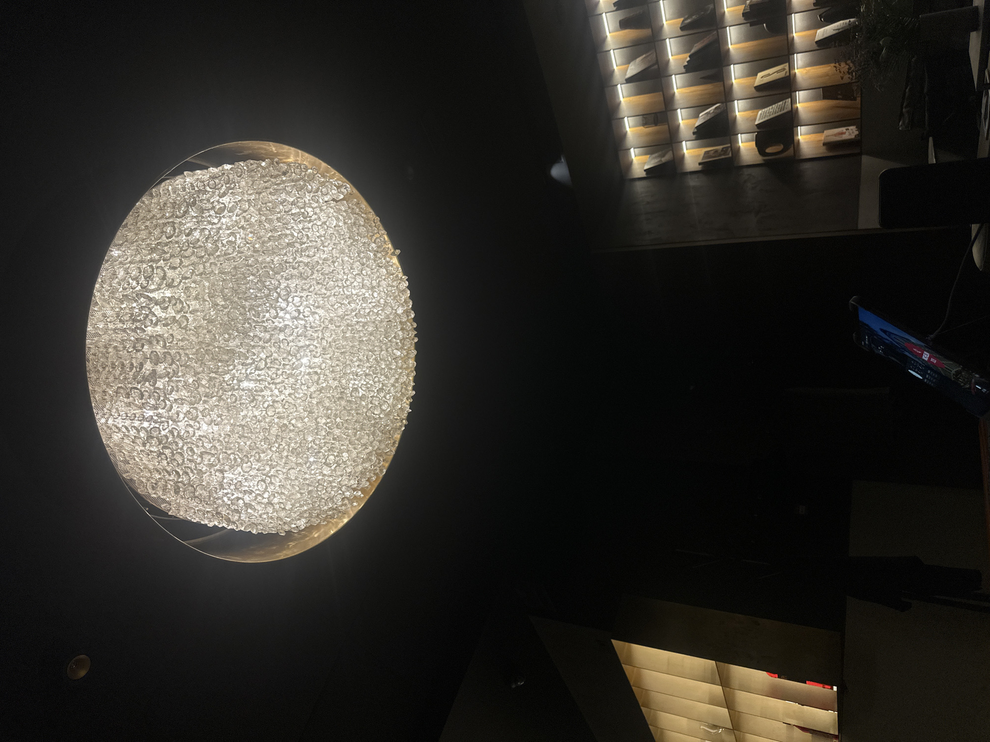 The image is taken indoors, likely in a restaurant or bar setting. A large, spherical chandelier dominates the left side of the frame, filled with many small, reflective or crystal-like elements that scatter the light. The ceiling and surrounding areas are dark, creating a high-contrast effect. In the background, towards the right side of the frame, is a wall with decorative lighting and geometric or rectangular elements. At the bottom right, a glimpse of another lit area is visible. Based on visual cues alone, it is not possible to determine the exact location in Sofia, Bulgaria, without further information or contextual clues.