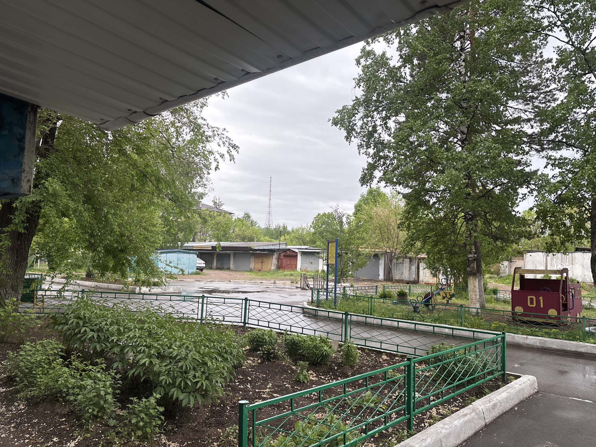 The image presents an overcast outdoor scene, possibly a residential or communal area, viewed from under a gray corrugated metal awning or roof structure. A cracked gutter is visible along the edge of this overhang. The ground, primarily dark soil and wet asphalt, suggests recent rain.
In the foreground, lush green bushes are planted behind a green metal fence with a diamond pattern. This fence extends into the midground, bordering a paved path and a wider asphalt area. To the right, within a fenced green space, are children's play structures: a red, train-shaped play toy with the yellow number "01" on its side, and a yellow and black spring rider, possibly in the shape of an animal like a bee.
Various deciduous trees with abundant green foliage, including a prominent birch, frame the scene. In the background, a row of garages with concrete walls and varied-colored doors (gray, red, yellow) lines a wider paved area. A light blue container-like structure is also visible near the left garages. A tall, slender antenna or transmission tower can be seen piercing the cloudy sky in the distance, slightly to the left of the center. A blue sign post is present in the midground, but any text on it is illegible. No people are visible in the scene. The overall atmosphere is quiet and subdued under the cloudy daytime sky.