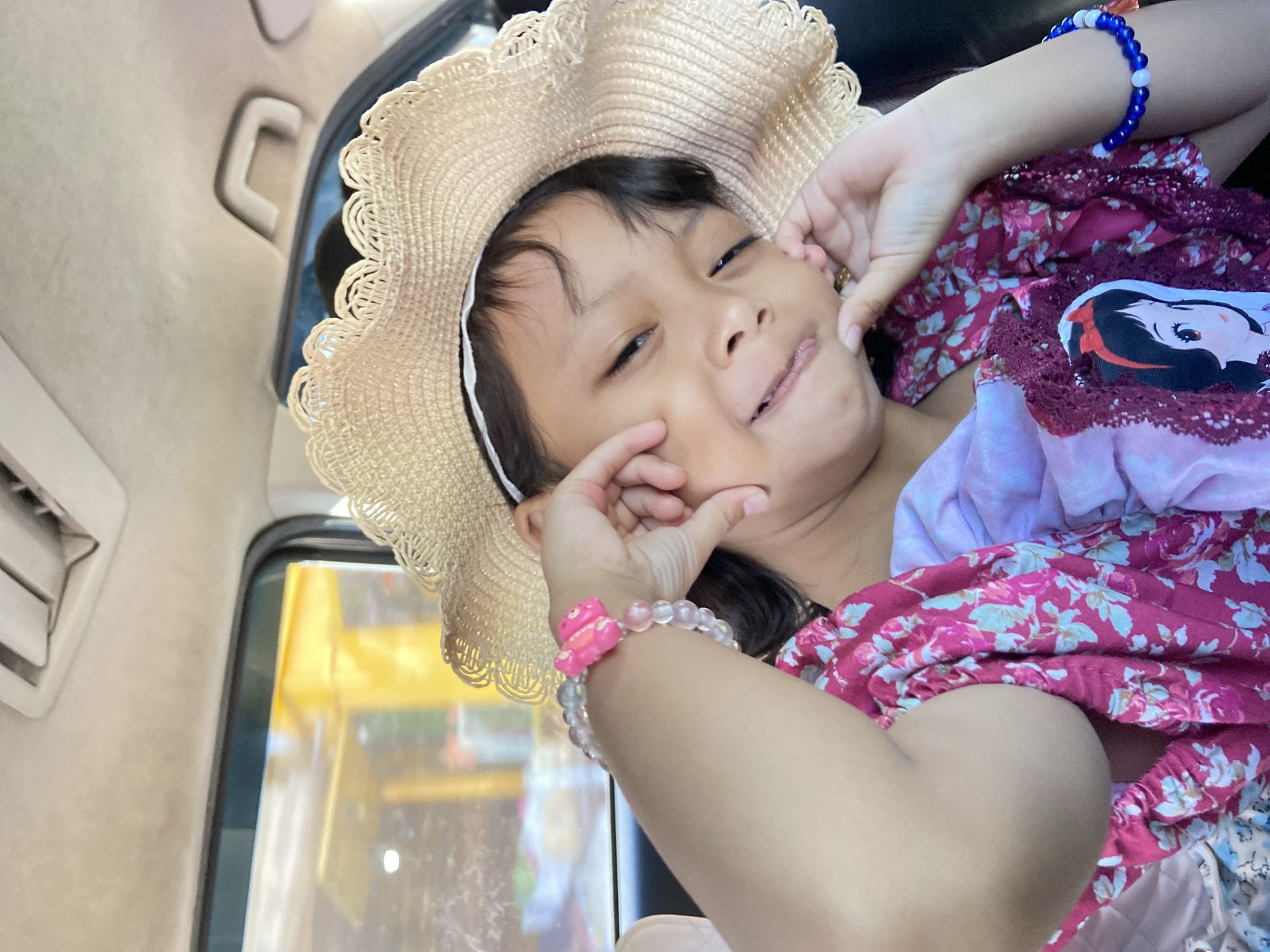 A young girl with dark hair wears a straw hat with a white trim and a pink floral-patterned dress with a cartoon character on the bodice. She has two beaded bracelets, one pink and one blue. She is inside a vehicle, likely a car, based on the interior elements visible in the background. The environment appears to be outside the car, as seen through the window. The location is Muktisari, Indonesia. The girl is holding her cheeks with her hands, making a funny face.