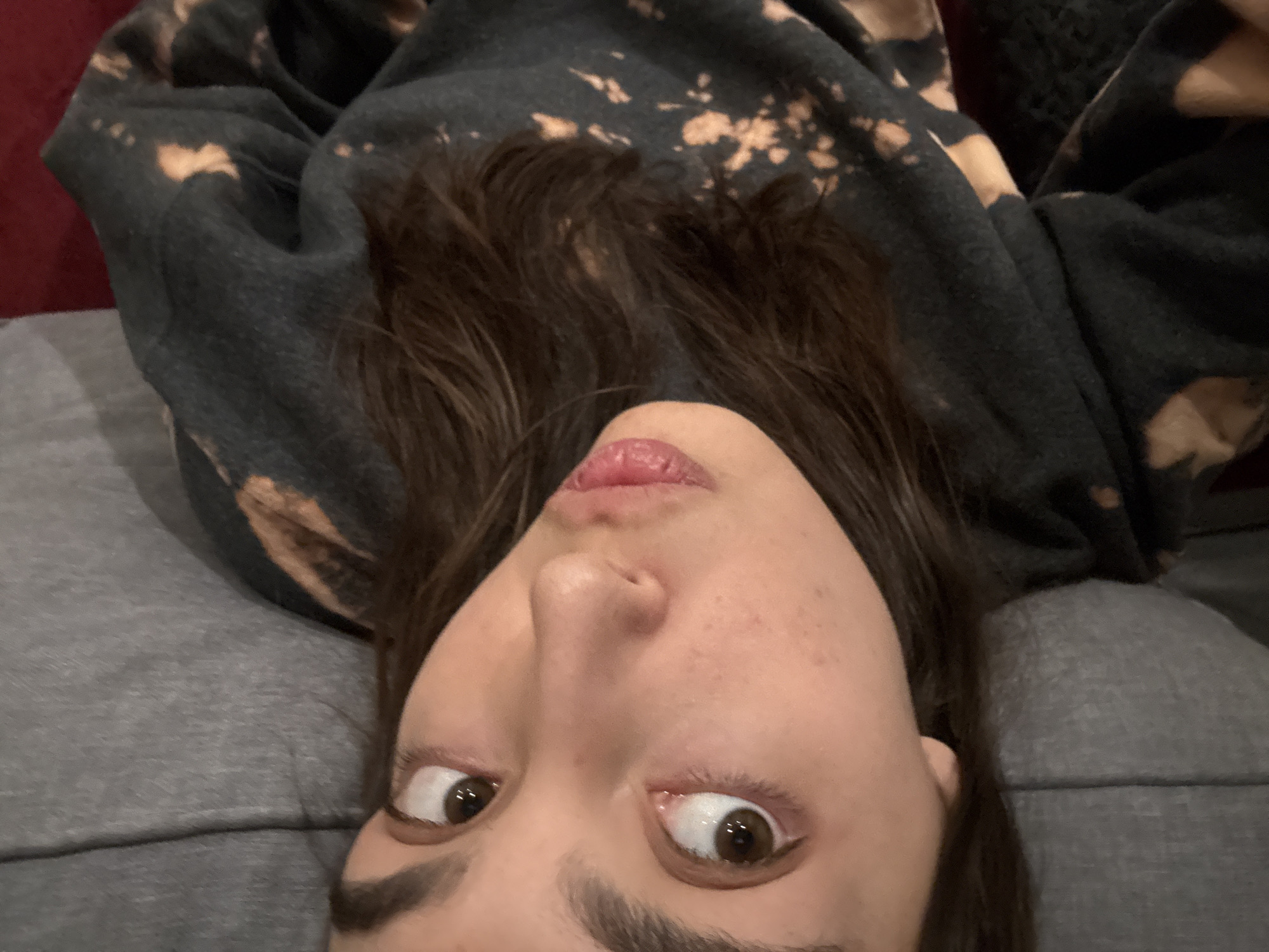 A young woman lies on a gray upholstered surface, presumably a couch or bed. She is wearing a dark gray hoodie with bleach spots. Her hair is brown and wavy. Her eyes are brown, and her gaze is directed upwards. The background is indistinct, with a glimpse of red upholstery in the upper left corner. The image is taken from a low angle, as if the camera is resting on the surface where she is lying. The scene appears to be indoors, but there are no distinct features to identify the location as Mexico City.