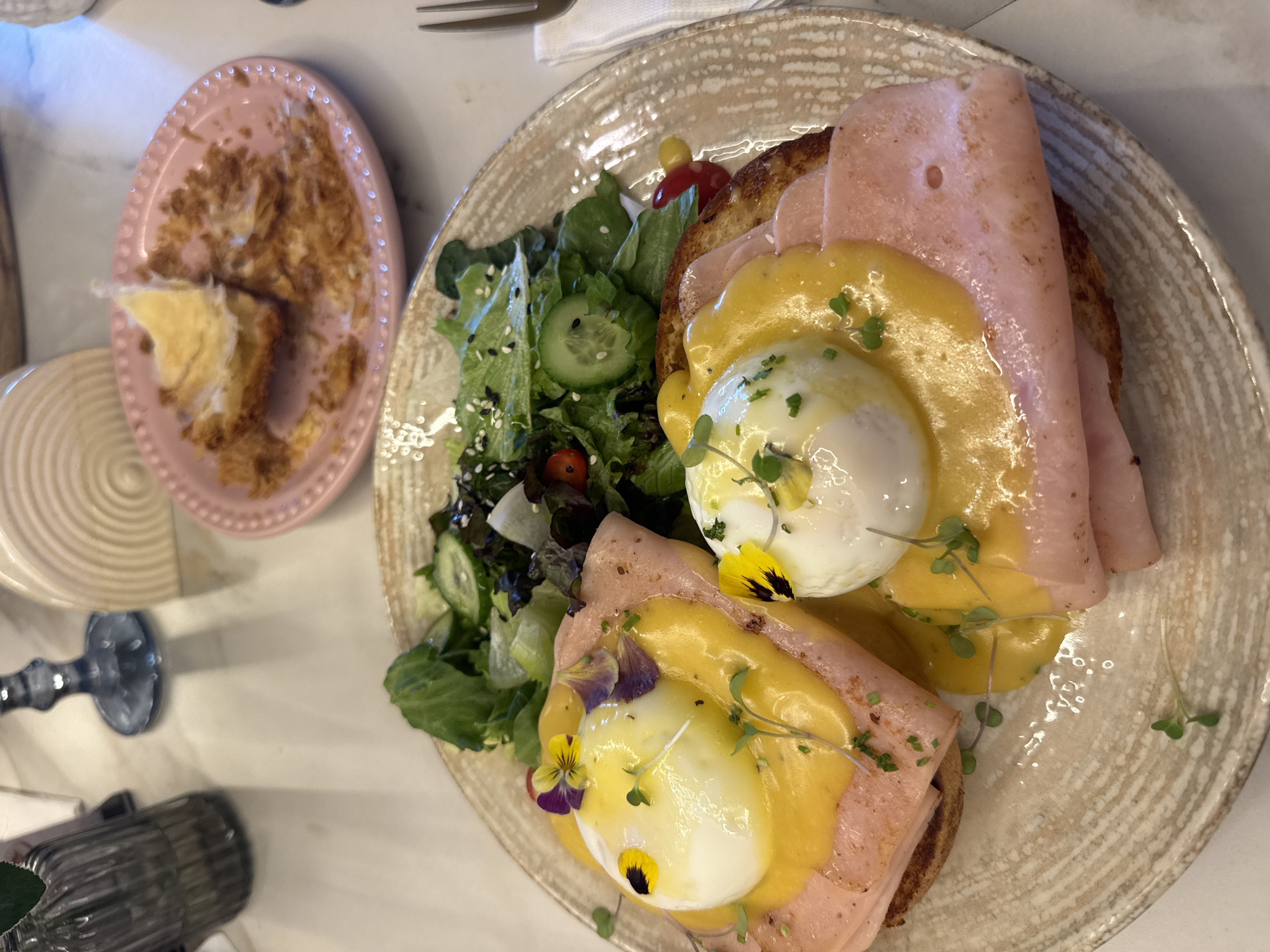 The image captures a table setting with a plate of Eggs Benedict, a side salad, and a dessert on a separate pink plate. The Eggs Benedict consists of two toast slices topped with ham, poached eggs, and hollandaise sauce, garnished with microgreens and small edible flowers. The side salad includes mixed greens, cucumbers, and cherry tomatoes, dressed with a vinaigrette and sprinkled with sesame seeds. The pink plate holds remnants of a pastry or cake. In the lower-left corner, a blue glass object suggests a vase or decorative item. The scene likely takes place in a restaurant or café in Puebla, Mexico.