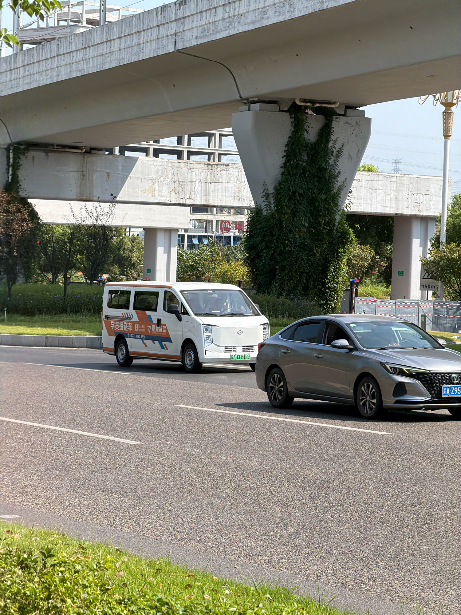 The image depicts a street scene in Jiangjin, China, under a large concrete overpass. A white van with orange and blue decals, likely a shuttle service, is driving in the left lane. The decals on the van feature Chinese characters, a phone number "(023) 4722 9898," and logos. In the right lane, a silver sedan is also driving. The sedan has a license plate that reads "A Z95" and appears to be an electric or hybrid vehicle due to its design. The road is paved and marked with white lines. Green grass and bushes are visible on the side of the road, and trees are scattered in the background. The overpass is a dominant feature, casting shadows on the road and supporting structures covered in green vines. The sky is clear and blue.