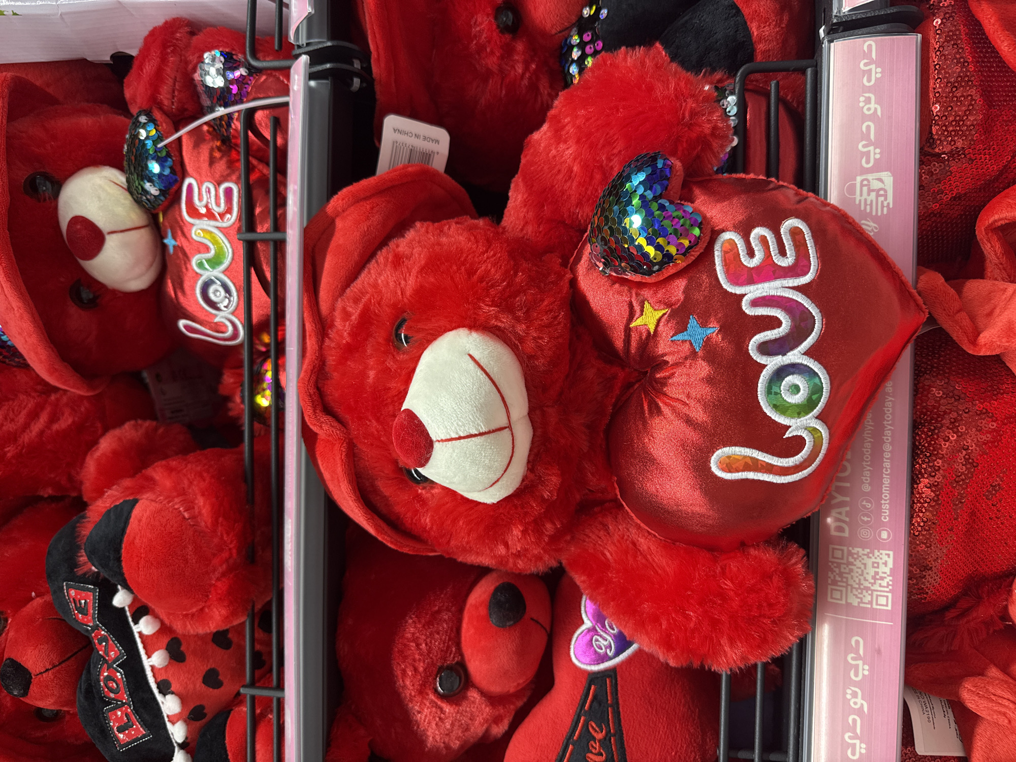 The image shows a close-up view of several red plush teddy bears displayed on shelves. The teddy bears are decorated with hearts and the word "LOVE" embroidered with colorful sequins. One teddy bear in the foreground is holding a red heart-shaped pillow with the word "LOVE" written on it in rainbow colors, and a small sequined heart. Another teddy bear to the left is wearing a black and red hat and has a black ribbon with the word "LOVE" on it. The shelves are made of metal and have pink labels with Arabic text and a QR code. The scene appears to be a retail display, likely in a shop or market in Deira, United Arab Emirates, given the Arabic text on the labels. There are no people visible in the image.