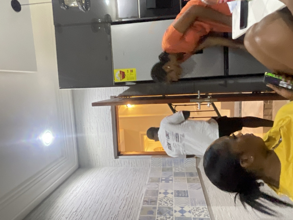 This indoor photograph, taken in Adenta Municipality, Ghana, captures a casual scene involving three individuals within a home environment. The image is rotated 90 degrees clockwise.
In the center, a woman dressed in an orange top is bending forward, appearing to interact with or look into the lower section of a dark grey or black refrigerator. A yellow energy efficiency label, featuring red text and graphics, is prominently displayed on the refrigerator door. To the left, a bright, circular ceiling light fixture emits a strong glow, illuminating the space.
In the middle ground, a man wearing a white t-shirt and dark bottoms stands with his back to the camera, positioned within a wooden-framed doorway or window opening that leads into another brightly lit room. The walls throughout the visible space exhibit a textured finish.
In the foreground, on the right, a second woman in a yellow top is partially visible, holding a mobile phone or similar device in her hand. The floor includes a section of patterned tiles featuring blue and white geometric designs, alongside a lighter, textured floor area. The atmosphere appears relaxed and domestic. No specific emotions are clearly discernible from the visible faces. Text on the man's white t-shirt is present but illegible.