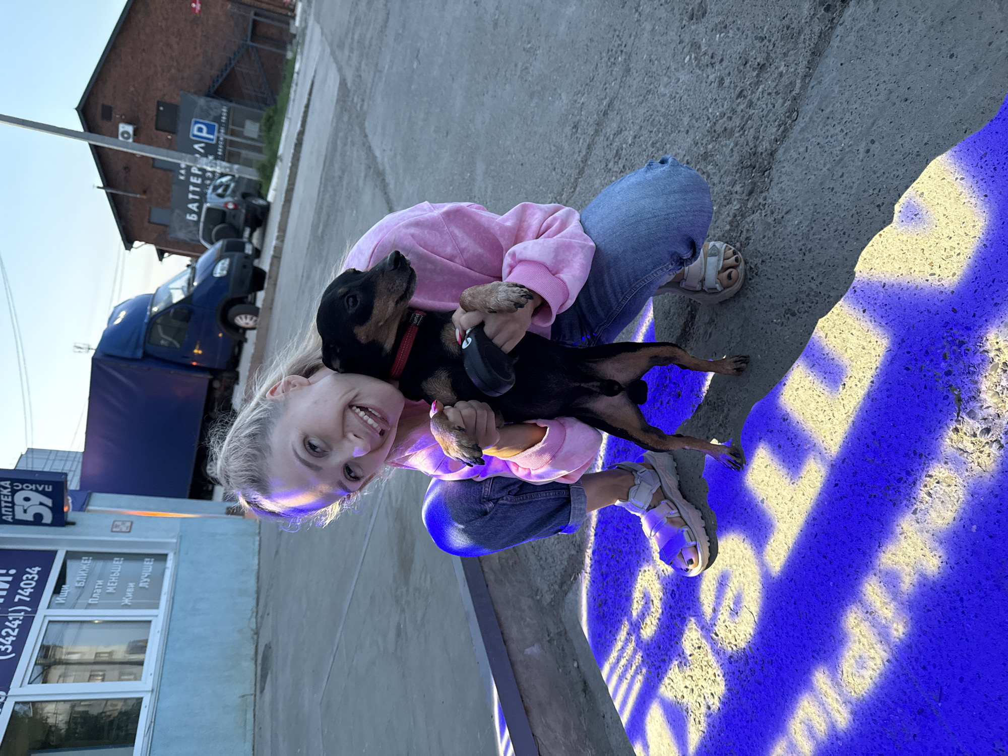 A woman is crouching and holding a small dog in an outdoor setting. She is smiling and wearing a pink sweatshirt and blue jeans, while the dog has a black coat with brown accents and a red collar. The background includes a building with signage indicating "Apteka 59" (Pharmacy 59), and a building with a sign for "BATTER A" and a parking symbol, suggesting this may be Chaikovsky, Russia. A blue van is also visible in the background. The ground has a blue and yellow pattern projected onto it.