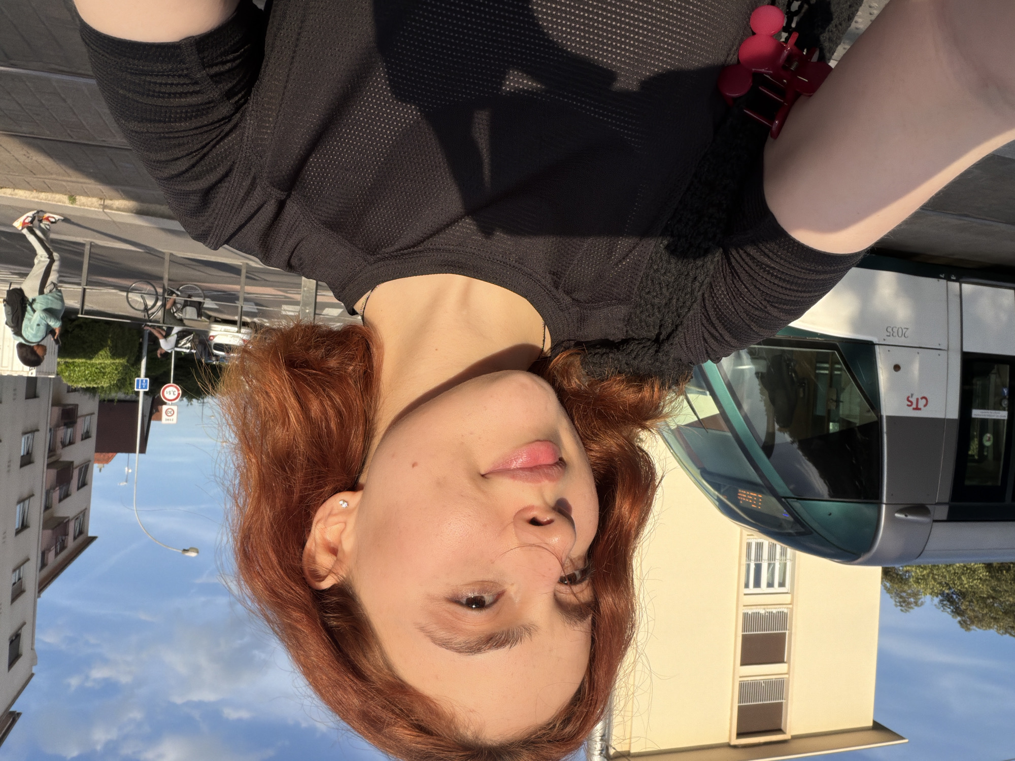 The image is a unique, upside-down selfie taken outdoors, featuring a young person with the urban environment appearing inverted above them.
The primary subject is a person with fair skin, dark eyes, and reddish-brown, medium-length hair. They are wearing a black, somewhat sheer, long-sleeved top. A small, sparkling stud earring is visible in their right ear (appearing on the left side of the frame). A pink, claw-style hair clip is tucked near their left shoulder. Strong sunlight illuminates their face, casting shadows.
The background depicts a city street on a sunny day. A blue sky with white clouds is visible. Buildings include a multi-story residential building with windows and balconies on the left, and another light-colored building on the right. A modern tram or bus, white and grey with green accents, occupies a significant portion of the right background, displaying "CTS" and the number "2035" on its side.
On the street, a sidewalk and road are visible. A metal railing separates the pedestrian area from what appears to be a track or median. Other people are in the background; one person with a backpack is walking away from the camera, and another is on a bicycle. Street furniture includes a streetlight pole and various road signs. Visible signs include a blue rectangular sign with white symbols, a circular speed limit sign with "50", and another circular sign displaying "3°C".
The activity is a casual outdoor selfie. The bright lighting and clear sky indicate daytime. The inverted perspective creates an unusual and dynamic composition. The architecture and the "CTS" text on the tram suggest a European city, specifically Strasbourg, France, as indicated by the context.