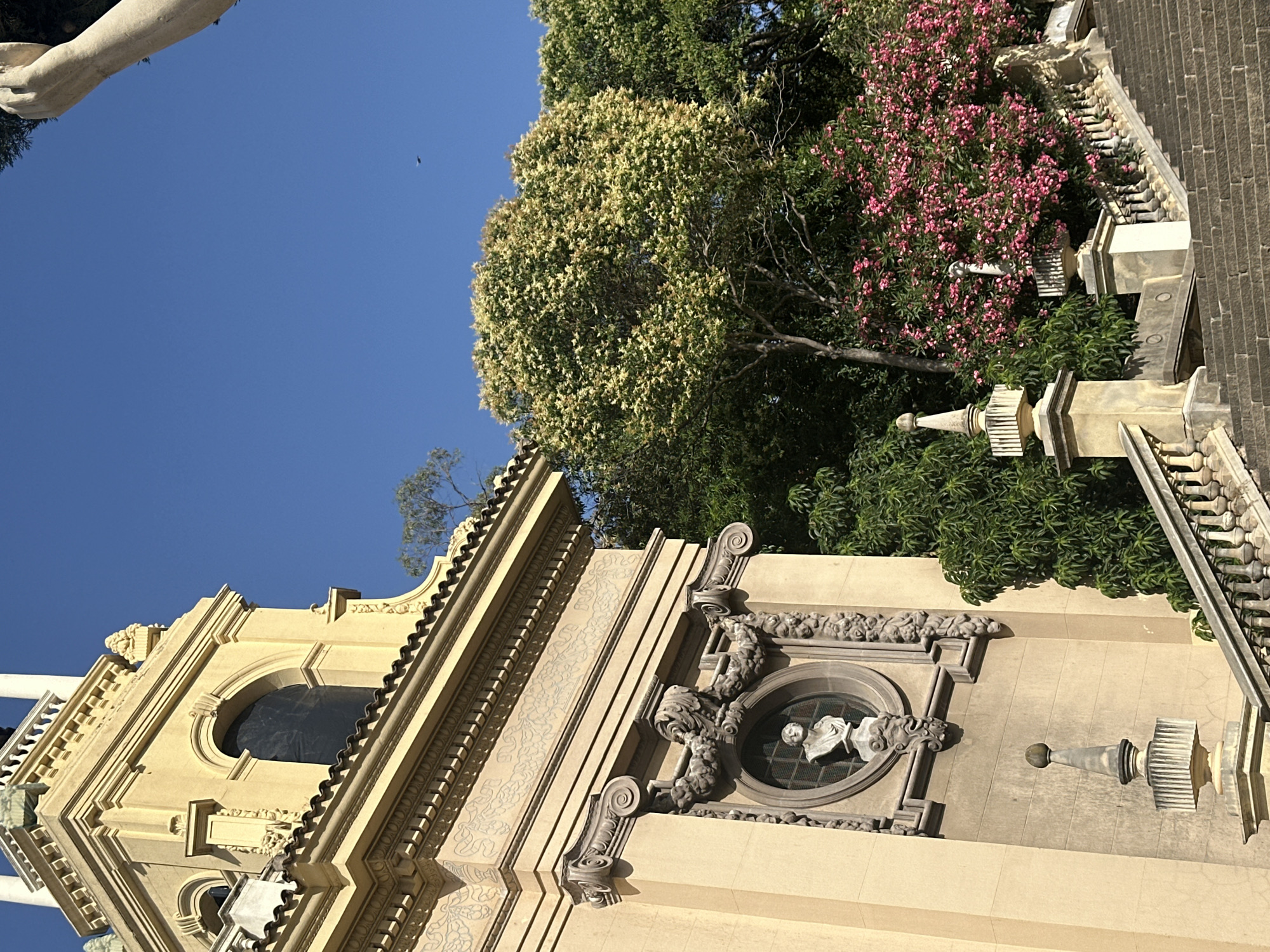 The image shows a close-up view of the exterior of a building in Barcelona, Spain, with a clear blue sky. The building is a light yellow color with ornate architectural details, including a corniced roof, decorative friezes, and scrolled corbels. A circular window features a bust of a person. In the foreground, a stone staircase with decorative balustrades ascends towards the building. Lush green trees and a vibrant pink flowering bush are visible behind the staircase and to the right of the building. A stone statue's arm is visible in the upper right corner of the frame. A tiny dark object, possibly a bird or insect, is visible against the blue sky in the upper middle section of the image. The scene appears to be a sunny day, likely in a park or a historic site within the city.