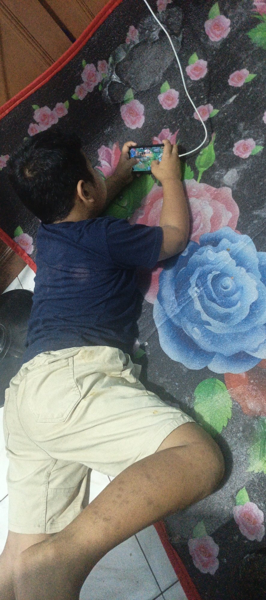 A young boy, viewed from above, lies on his stomach on a dark, patterned mat adorned with large floral designs in shades of pink, blue, and red. He is wearing a navy blue t-shirt and light beige shorts. The boy is holding a smartphone horizontally with both hands, engrossed in playing a game that is visible on the screen. A white charging cable is connected to the phone and trails upwards out of frame.
The setting appears to be indoors, possibly a bedroom or living area, suggested by the presence of a wooden surface on the left and a tiled floor visible beneath the mat and the boy's lower legs. The lighting is somewhat dim, implying it could be evening or a room with limited natural light. The boy's posture and focus on the game indicate he is occupied and engaged in recreational activity. His skin has a warm tone, and there are some light markings on his shins. The overall scene captures a moment of quiet leisure.