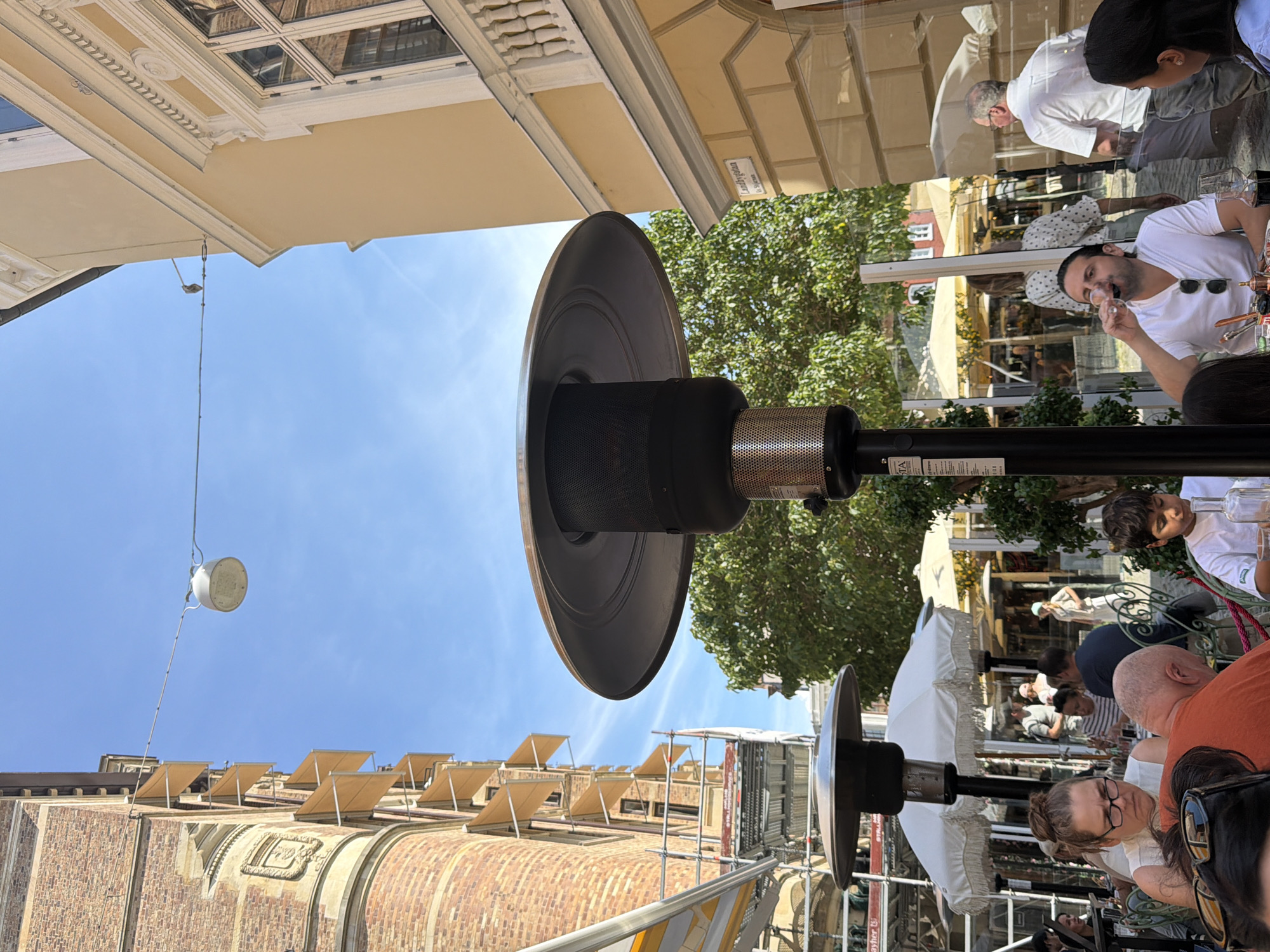 The image depicts an outdoor dining scene on a sunny day. In the foreground, a man in a white t-shirt drinks from a wine glass. To his left, a woman with glasses looks down at her table. Behind them, several other diners are visible at tables with umbrellas and patio heaters. The background shows buildings with brick and yellow facades, as well as trees and scaffolding. A street sign that reads "Landbygatan" is visible on the yellow building, indicating the location is Landbygatan in Malmö, Sweden.