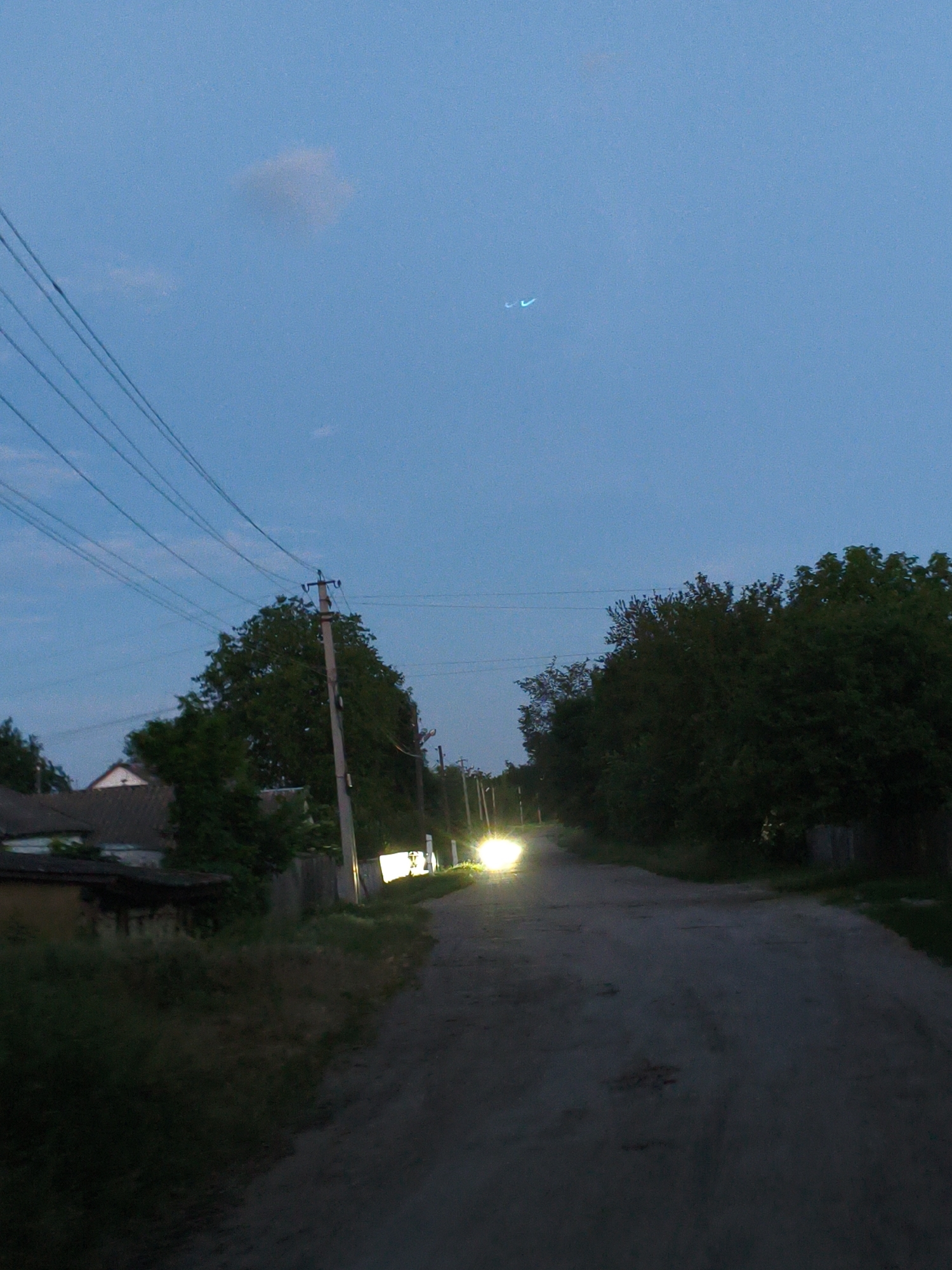 This is a low-light, eye-level, outdoor photograph taken at dusk or dawn on a rural dirt road in Ustyvytsia, Ukraine. The sky is a pale blue with a faint cloud formation in the upper left quadrant and two faint, curved, light blue streaks in the upper center. A cluster of power lines runs diagonally from the upper left to the center of the frame.
On the left side of the road, a small, single-story building with a dark roof is visible. Adjacent to it is a wooden fence and lush green vegetation. Further down the road on the left, a utility pole with wires is prominent.
The road itself is unpaved and dark, with some debris or unevenness visible. Trees with dark green foliage line the right side of the road, creating a dense wall of vegetation that obscures any structures behind them.
Several utility poles with overhead wires are spaced along the right side of the road, receding into the distance. The headlights of an approaching vehicle are visible at the far end of the road, casting a bright white light that illuminates the immediate vicinity of the road surface. The overall impression is one of a quiet, rural setting as daylight fades.