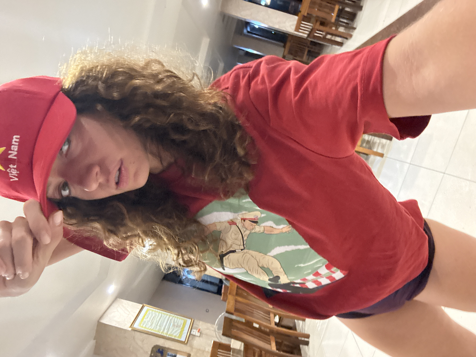 A selfie of a person with curly brown hair wearing a red cap with "Việt Nam" in white lettering and a red t-shirt with an image of a man in a tan uniform surfing, taken indoors. The person is holding the brim of the cap. The background includes tile flooring, wooden chairs and tables, and a picture frame on the wall. The location is inferred to be in Tú Lệ, Vietnam, based on the cap's lettering.