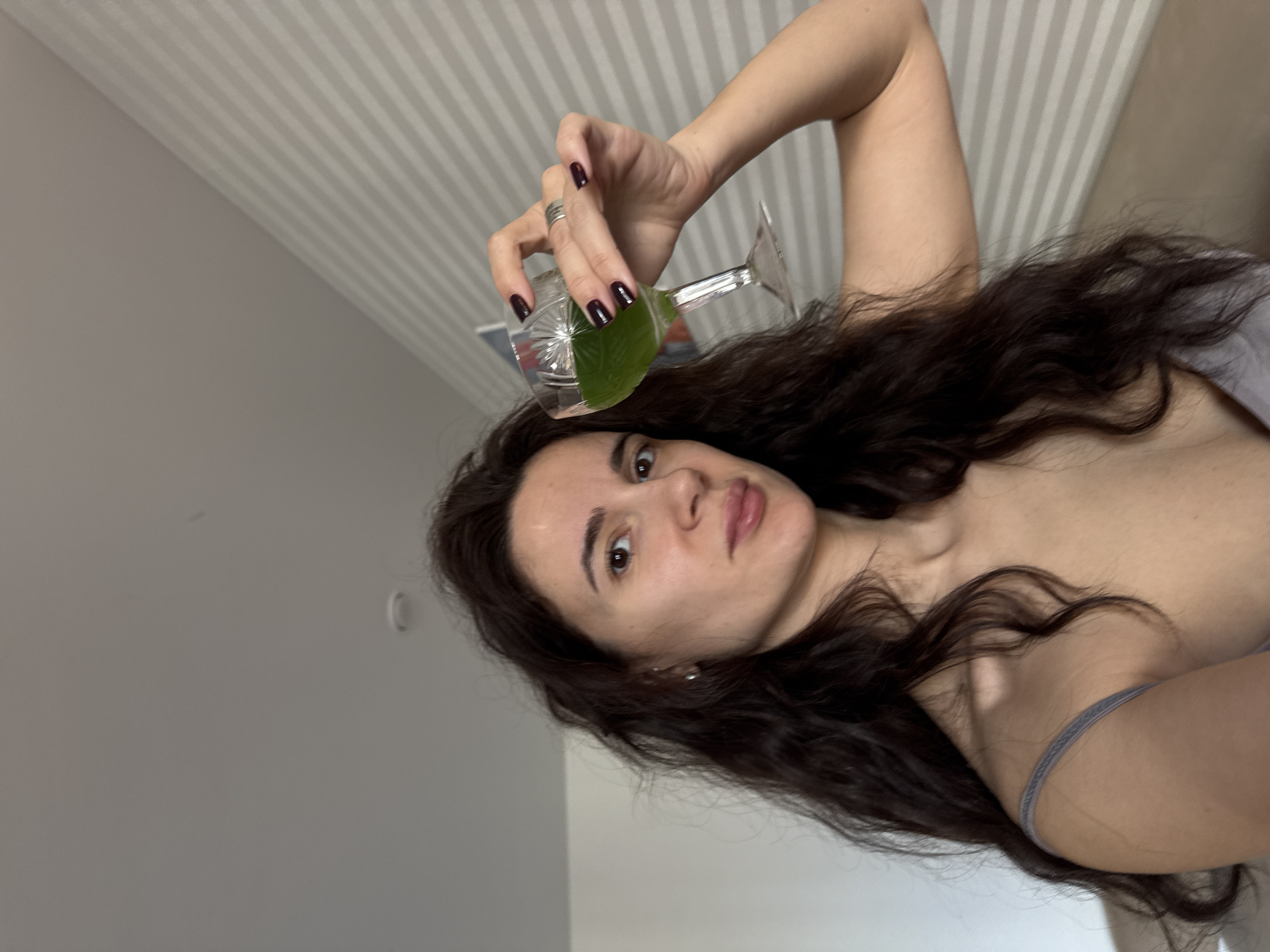 A woman with dark hair and a fair complexion is holding a glass of green liquid. She has dark nail polish and is wearing a ring. The background shows a wall with vertical stripes. The environment appears to be indoors. There is no indication of the location being Chaikovsky, Russia.