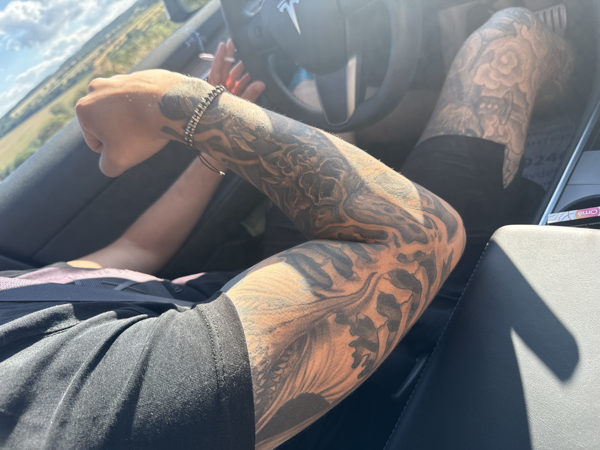 The image shows a close-up of a person's arms and legs, heavily tattooed, inside a car. The person is wearing a black t-shirt and black shorts. Their left arm, covered in a sleeve tattoo, rests on their lap. Their right arm is on the steering wheel, which has a Tesla logo. A bracelet is visible on their right wrist. In the background, through the car window, a rural landscape with fields and a cloudy sky is visible. The sun is shining, casting shadows inside the car. The specific location of Yabalchevo, Bulgaria is not visually discernible from the image itself.