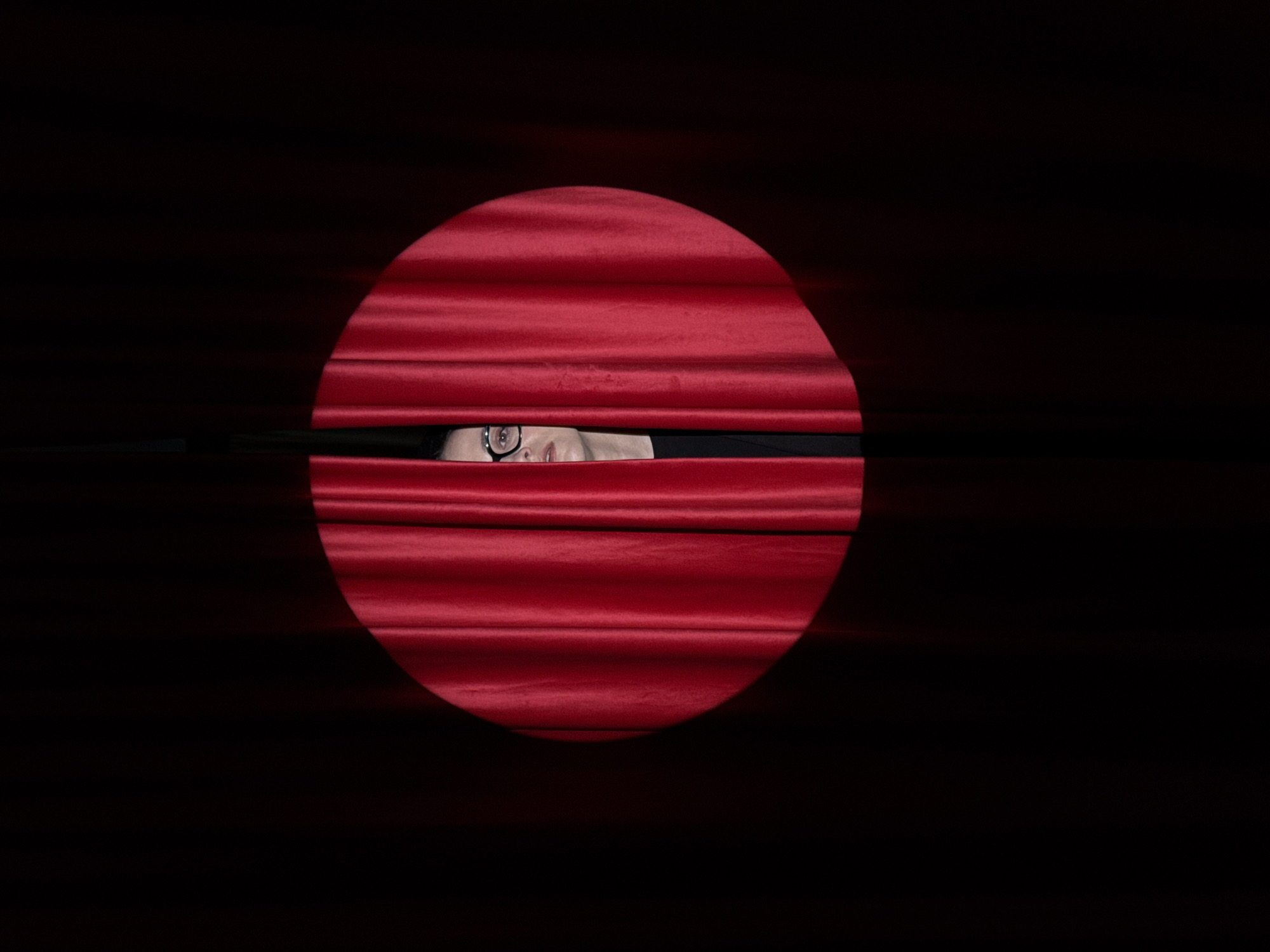 A person with glasses peers through a narrow opening in horizontally-oriented, dark-red blinds within a dark, circular area against a black background. No specific details indicate the image location.