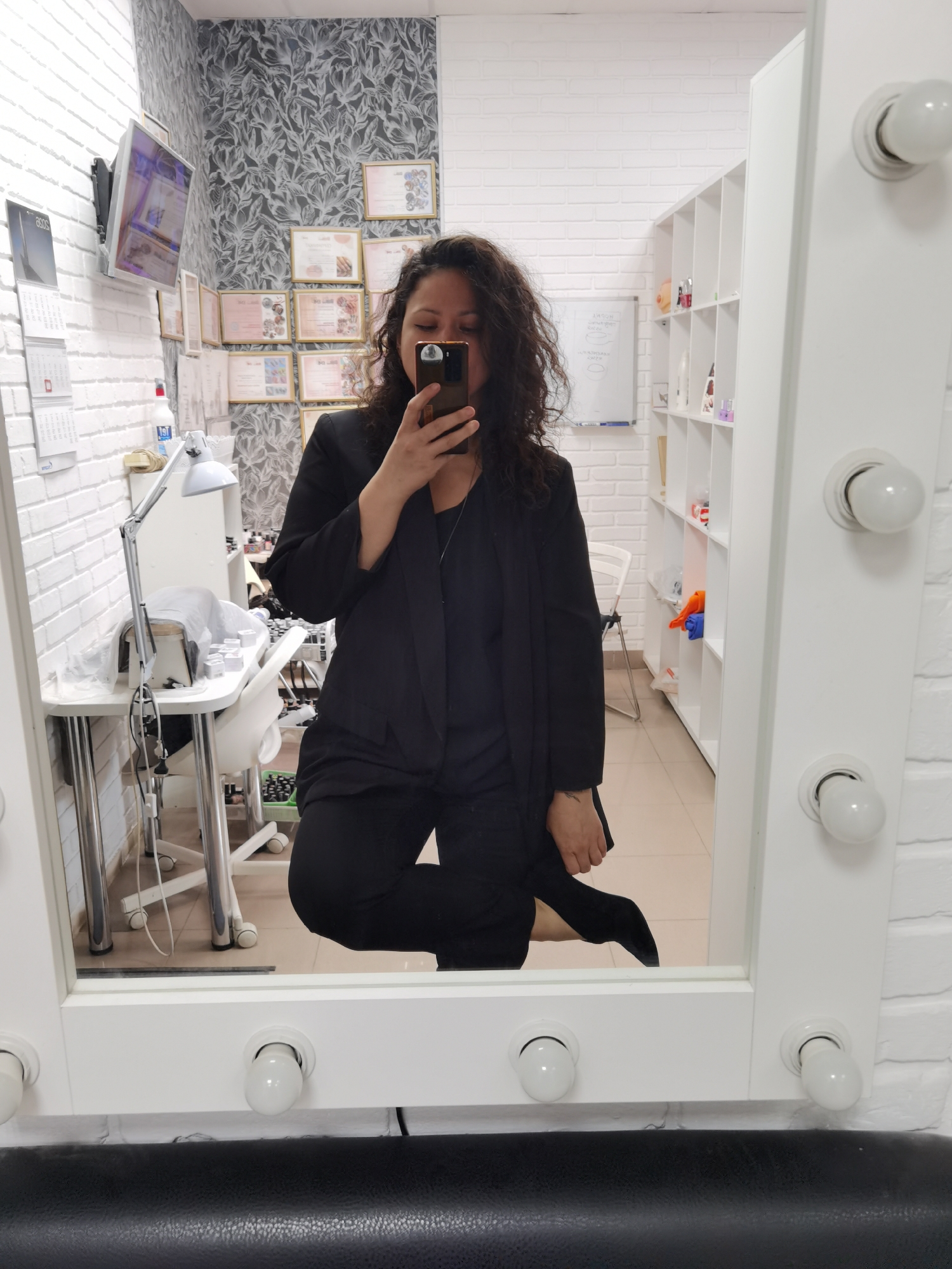 A woman stands in what appears to be a commercial or salon setting in Chaikovsky, Russia, taking a selfie in a mirror with light bulbs surrounding the frame. She wears a black blazer and pants, possibly a suit, and is looking at her phone. The location has white brick walls on two sides; one wall is decorated with framed certificates and a calendar. There is a white table with beauty supplies and a lamp, as well as a white shelving unit containing various items. The floor is a neutral color.