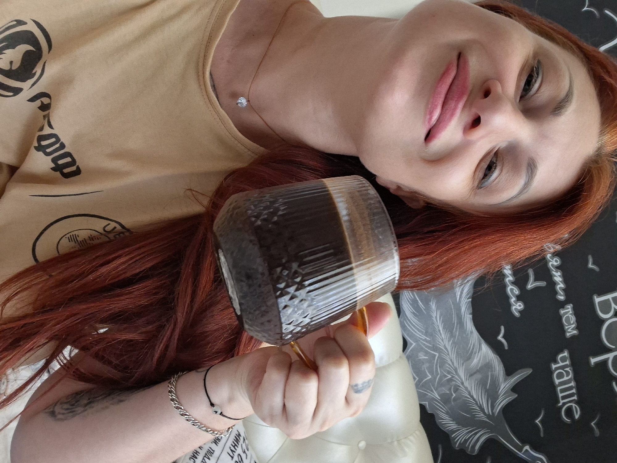 A woman with red hair and light skin is shown in a selfie, holding a glass mug of dark liquid. She is wearing a beige t-shirt with black graphics. A faint tattoo is visible on her right forearm. She is also wearing a thin necklace with a small, clear pendant. The background appears to be a wall with white chalk drawings of birds and text. This image was taken in the city of Myadzyel, Belarus.