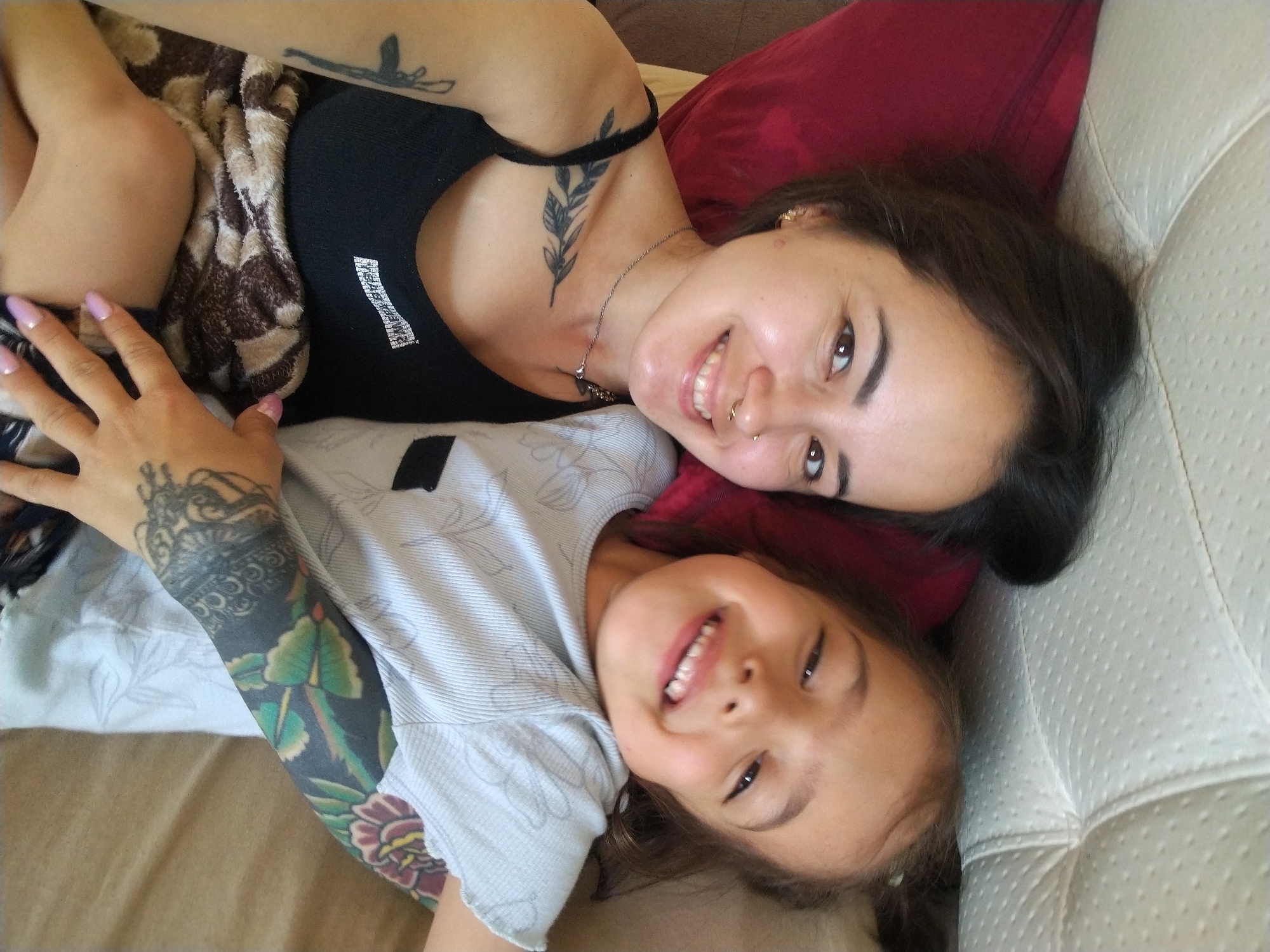 This media file features a close-up, overhead shot of two females, an adult and a child, lying down together. The adult, on top, has dark hair and is looking towards the camera with a smile, displaying a nose ring and a tattoo on her collarbone of a branch with leaves. She is wearing a black tank top. The child, positioned below, is looking directly at the camera with a wide, joyful smile, showing visible teeth. The child has dark hair and is wearing a light gray t-shirt. The adult's arm is visible, adorned with extensive tattoos on her forearm, including floral designs. They appear to be on a soft surface, possibly a bed or couch, with a textured, light-colored material visible on the right side of the frame, possibly upholstery. A red pillow is also visible. The overall impression is one of warmth and affection between the two subjects. The lighting suggests it is daytime. No text is clearly visible in the image.