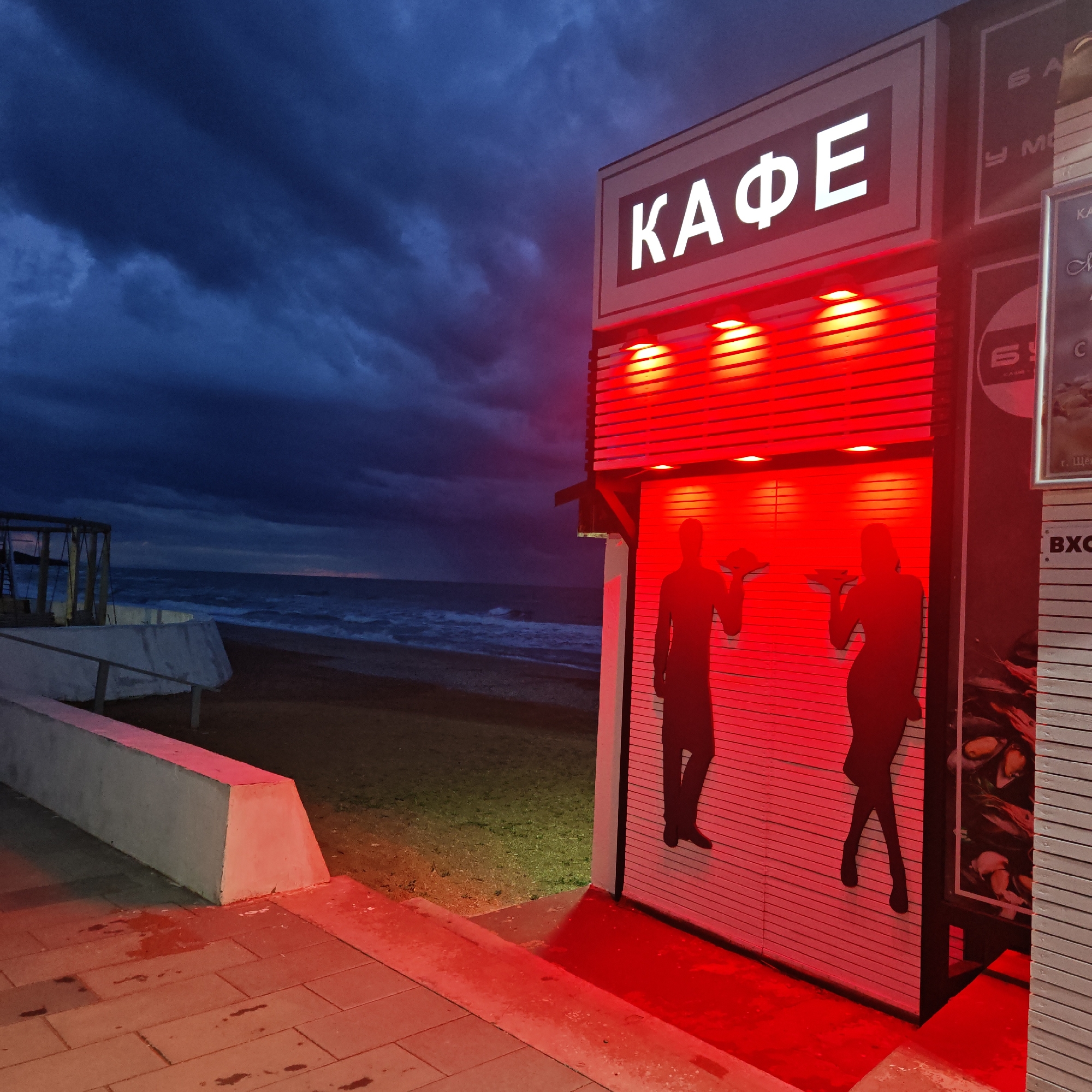 The image depicts a cafe, "KAFE" in cyrillic letters, illuminated with bright red lights, located near the beach in городское поселение Щелкино, Ukraine. The cafe's facade features silhouettes of a man and a woman holding trays. The scene is set under a stormy sky with dark clouds looming over the sea. In the background, the beach is visible, along with waves and a section of a white concrete barrier. The foreground shows a paved area, also illuminated with red light.