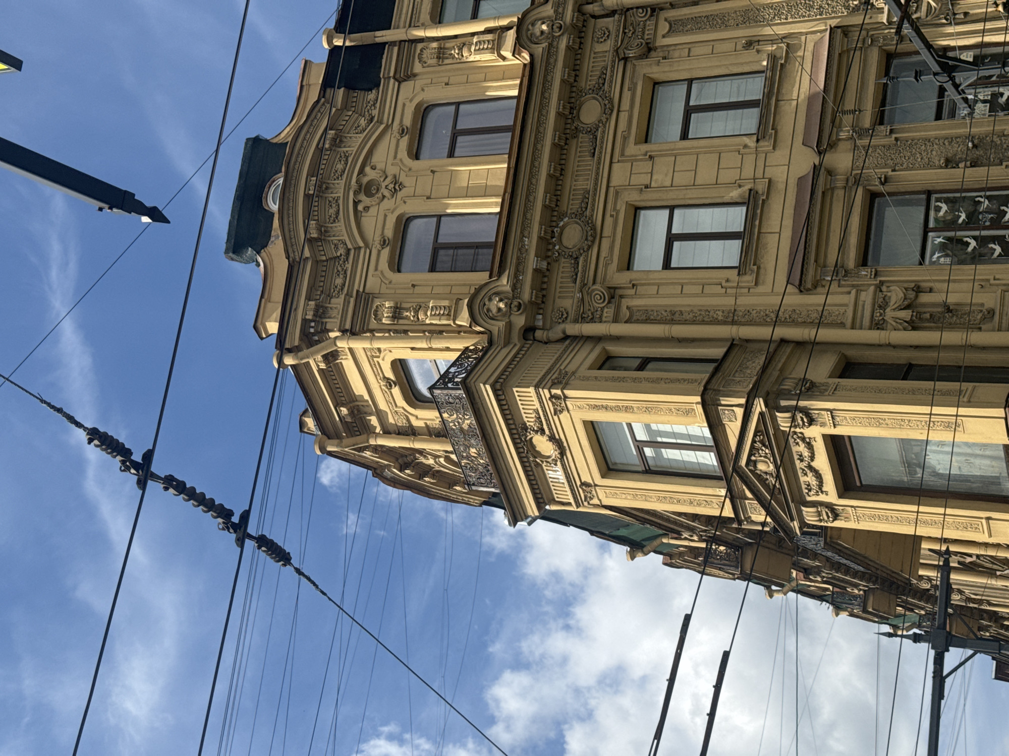A low-angle photograph captures a portion of an ornate, multi-story building against a clear to partly cloudy blue sky. The building, painted in a warm yellowish-brown, displays a richly decorated facade with elements characteristic of historical European architecture, possibly Art Nouveau or Neo-Baroque. Visible details include intricately carved pilasters, cornices, and moldings featuring floral patterns, geometric designs, and what appear to be decorative masks. Several rectangular windows with dark frames are present, some reflecting the sky. A small wrought-iron balcony protrudes from one of the upper levels, and one window on the right side features a distinctive decorative insert, possibly stylized bird silhouettes.
Numerous black electrical or trolley wires crisscross the frame in the foreground, forming a dense network that partially obscures the sky and the building. A dark metal street lamp or pole is also visible, extending into the frame from the left side. The sky is predominantly blue with scattered white, wispy clouds, indicating daytime conditions. The perspective looks sharply upwards, emphasizing the building's height and the overhead urban infrastructure. The scene is static, with no people or specific activities taking place. There is no visible text. The architectural style is consistent with buildings found in Saint Petersburg, Russia.