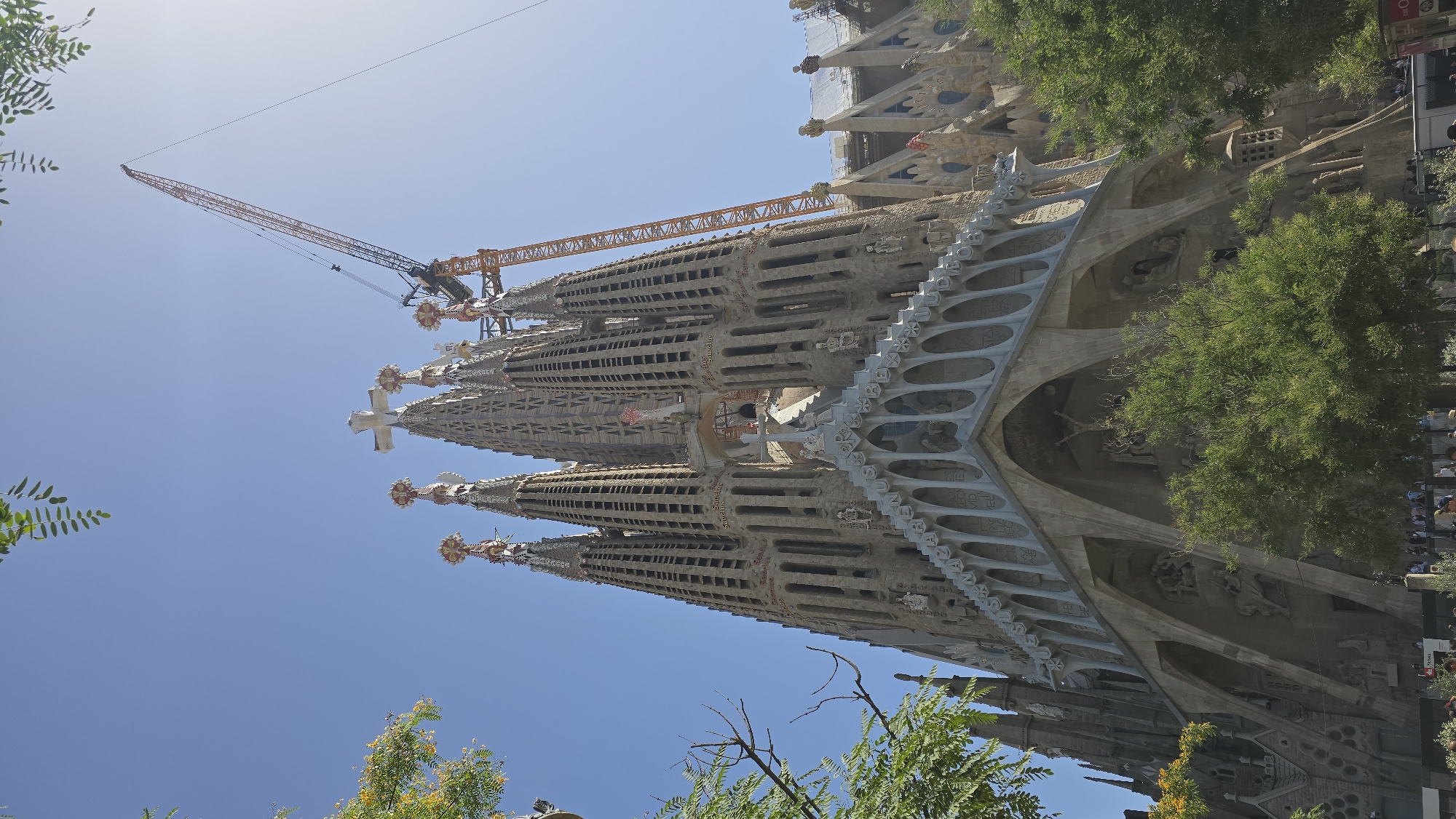 This is a low-angle, full shot of the Sagrada Familia in Barcelona, Spain, on a clear day. The iconic basilica is in the center and right of the frame, with its distinctive spires and detailed facade. A large orange construction crane is visible towering over the right side of the basilica, indicating ongoing construction or renovation. Lush green trees partially frame the bottom and left sides of the building, with some foliage also visible at the top. The sky is a bright, clear blue. At the bottom of the image, a crowd of people are gathered, appearing to be tourists or visitors, with some walking and others standing. The architecture is intricate, with many sculptures and architectural elements visible on the facade.