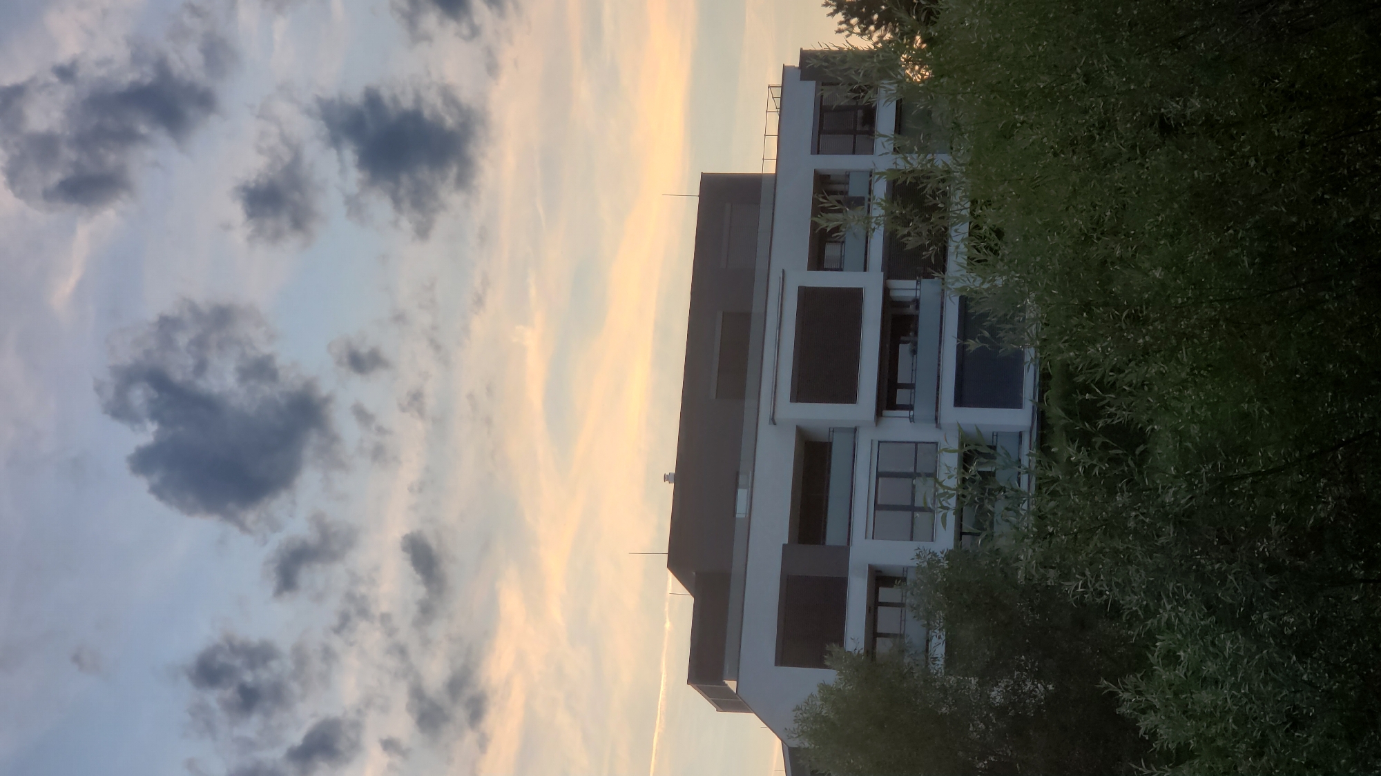 The image shows a modern apartment building with a light-colored facade and dark-tinted windows, partially obscured by lush green foliage in the foreground. The building features multiple stories with balconies and horizontal window elements. Above the building, the sky displays a mix of soft pastel colors, indicating either sunrise or sunset, with scattered dark clouds and a faint contrail visible. The scene appears to be an exterior view of a residential area, possibly on the outskirts of a city, given the presence of trees and greenery. No people or specific actions are depicted in the image. This image is not identifiable as being located in Prešov, Slovakia.