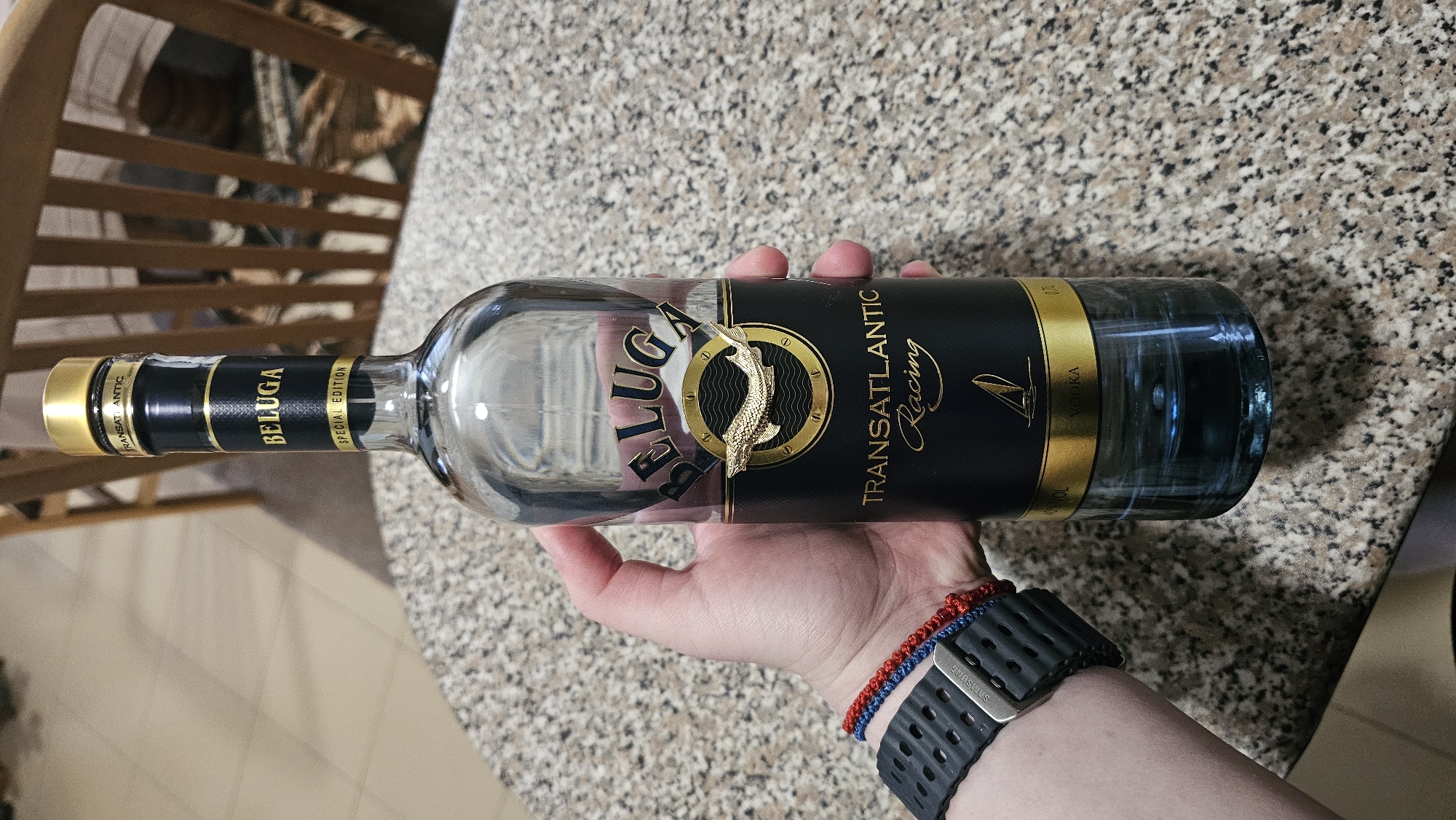 A hand holds a bottle of Beluga Transatlantic Racing vodka. The bottle is mostly clear but has a black label with gold accents and the brand name "Beluga" prominently displayed. The label also indicates that it's a "Special Edition." A gold-colored fish emblem is affixed to the front of the bottle. The background consists of a granite-look countertop, a portion of a wooden chair, and an arm with a black Samsung watch and two bracelets. The image does not show or suggest the location of Rogachevo, Bulgaria.