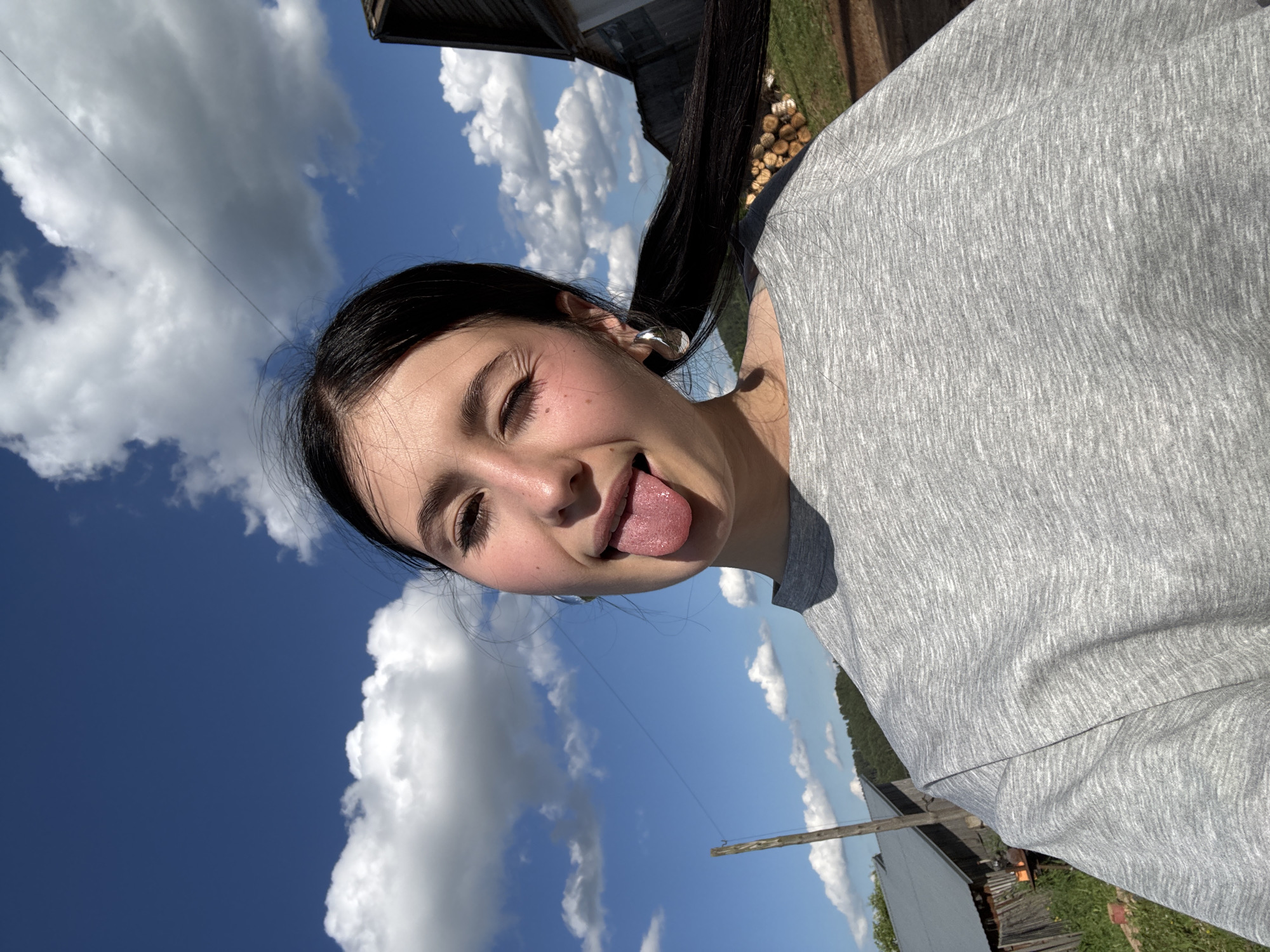 A young woman is taking a selfie outdoors on a sunny day. She has dark hair pulled back into a ponytail and is wearing a grey t-shirt. She is winking and sticking her tongue out at the camera. In the background, there is a blue sky with white clouds, some trees, and rustic buildings. The location is identified as the city of Fokinskoye, Russia.