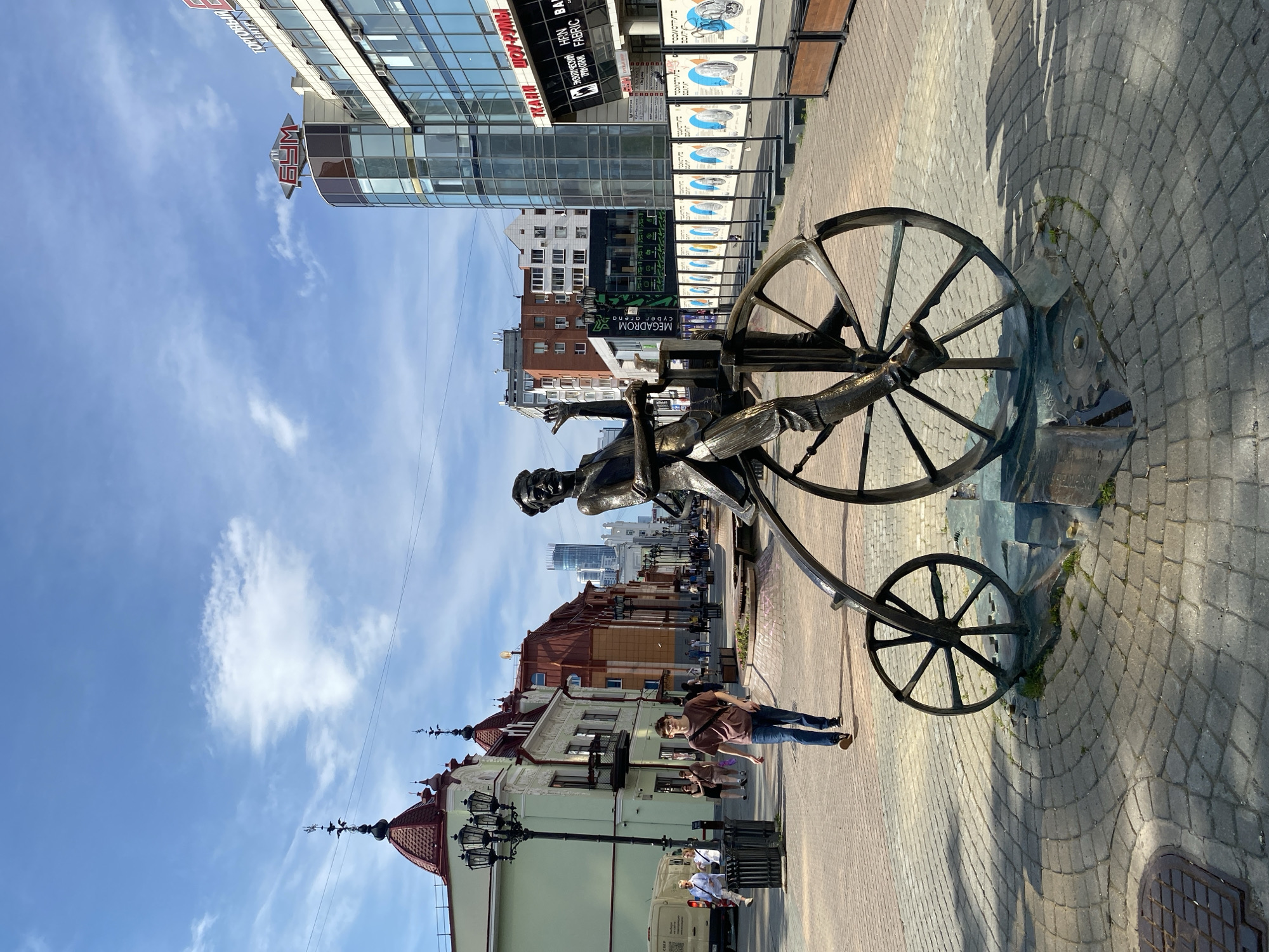 A statue of a man on a vintage bicycle is located on a cobblestone sidewalk in Yekaterinburg, Russia. The statue is made of bronze and depicts a man wearing a hat and a suit, with his hands on the handlebars of the bicycle. The bicycle has a large front wheel and a smaller back wheel. The statue is situated in front of a modern glass building with the word "Бум" in red lettering on top. The street is lined with buildings of various architectural styles, including older buildings with red roofs and a taller skyscraper in the distance. Several people are walking on the sidewalk, and a white van is parked on the left side of the street. The sky is blue with scattered clouds. The statue is a popular landmark in Yekaterinburg, and it is often photographed by tourists.