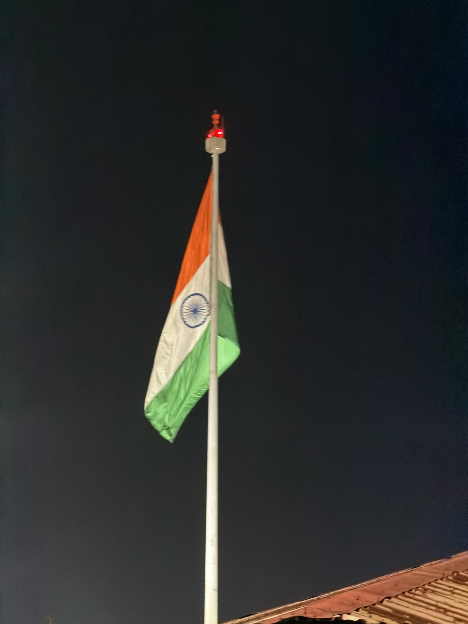 An Indian flag waves from a flagpole against a dark night sky. The flag has three horizontal bands of saffron, white, and green, with a navy blue Ashoka Chakra at its center. At the top of the flagpole, a red light is visible. In the lower right corner of the image, the corrugated rooftop of a building is partially visible. The background is a deep, uniform black, indicating nighttime. The image appears to be taken in Kochi, India, given the context provided.