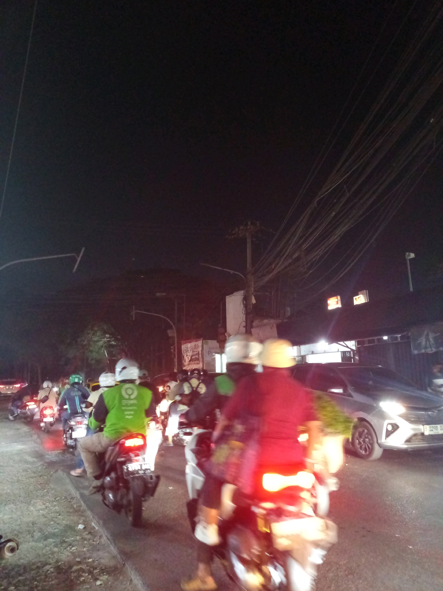 The image depicts a street scene at night, likely in Karangsatria, Indonesia, characterized by heavy traffic and a dark sky illuminated by artificial lights. Numerous motorcycles are present, some carrying passengers, with riders wearing helmets. Several motorcycles have illuminated taillights, indicating movement or stopped traffic. One motorcycle in the foreground has a license plate that is partially legible, showing "B 2709 TAM".
A silver car is visible in the middle ground, to the right of the frame, with its headlights on. Buildings are present on the right side of the street, with some illuminated windows and signage, one sign featuring the logo of "Gojek," a ride-hailing company. Overhead, a dense network of power lines crisscrosses the sky, a common sight in urban Indonesian environments. The overall impression is one of a busy, active street during nighttime hours, with a focus on motorcycle traffic.
