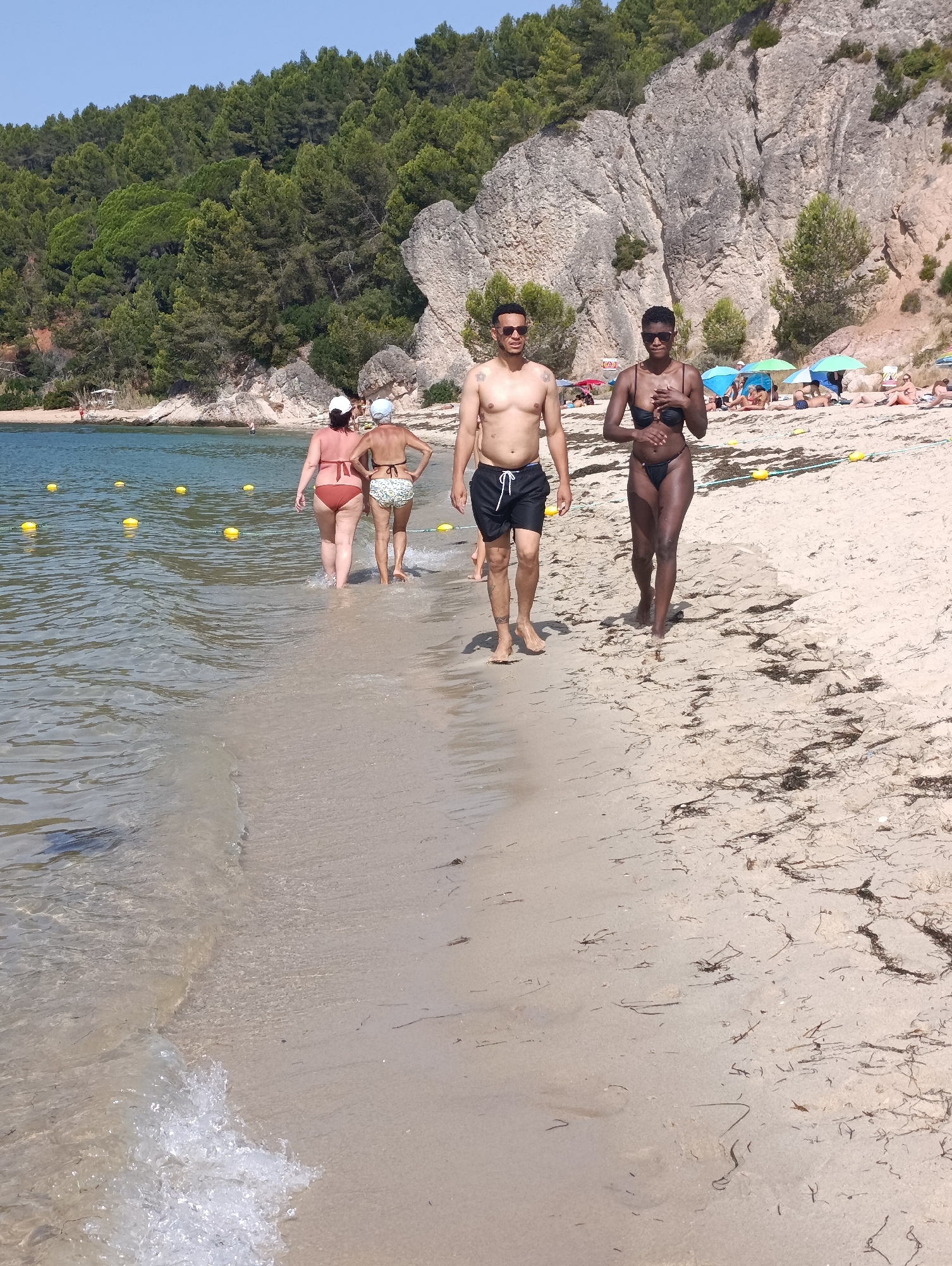 The image shows a sunny day at a beach in Setúbal, Portugal. In the foreground, a couple walks along the shoreline. The man, shirtless and wearing black swim trunks, walks on the sand, while the woman, in a black bikini, walks in the shallow water. Behind them, two women in swimwear also walk in the water, one with her back to the camera. The beach is lined with green trees and rocky cliffs in the background. Several beach umbrellas and sunbathers are visible further down the beach. Yellow buoys float in the water, marking a swimming area. The sand is light brown and appears to be mixed with seaweed. The water is clear and calm, with gentle waves lapping at the shore. The sky is a clear blue.