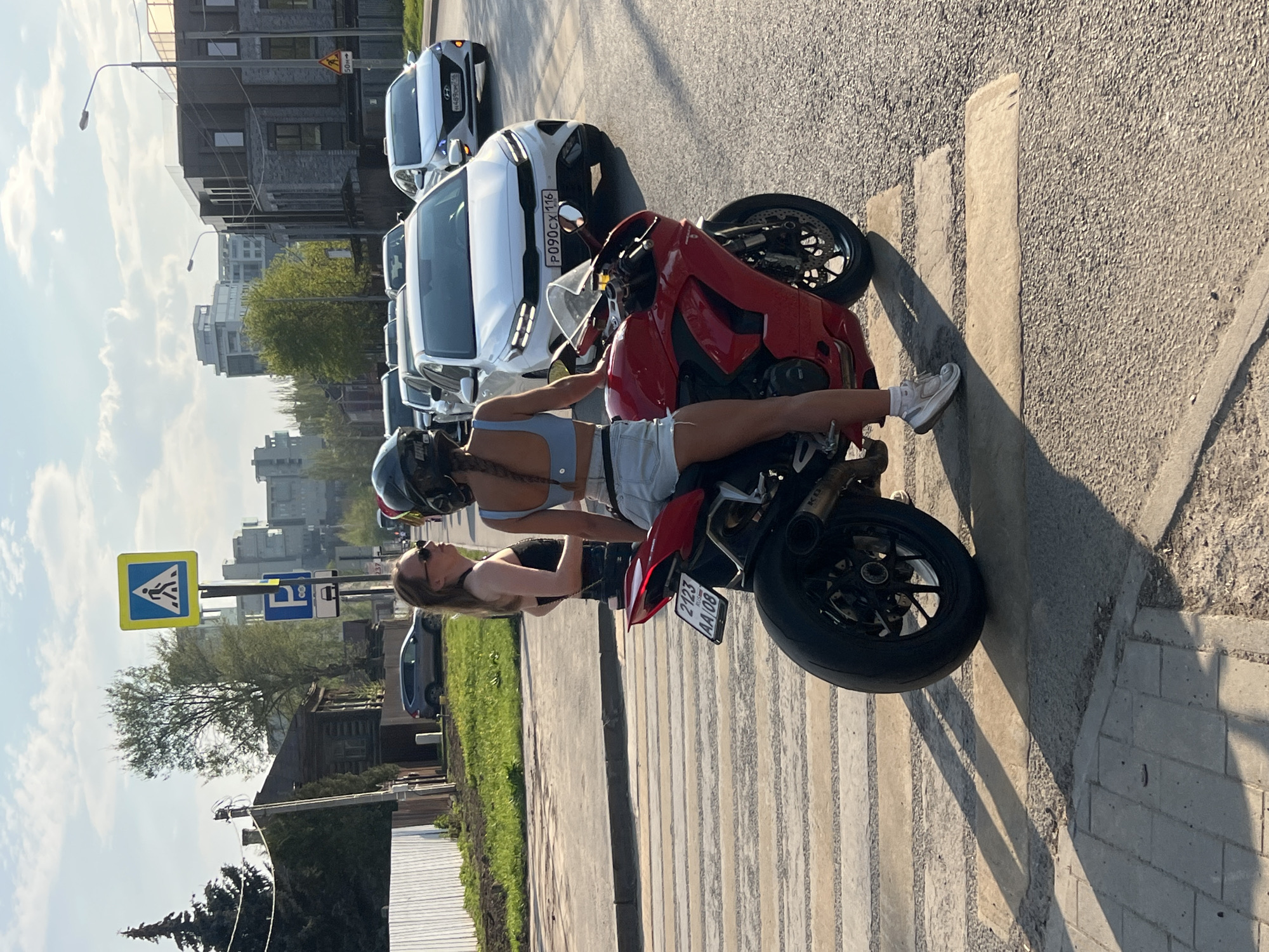 A color photo captures a woman lying across a red motorcycle on a city street in Kazan, Russia. She has light skin, a light blue top and white shorts, a helmet and white sneakers. The motorcycle is parked on a pedestrian crosswalk. Behind the motorcycle, several white cars are parked parallel to the crosswalk. Across the street, a green grassy area separates the sidewalk from a low, white fence. Further back, there is a mix of modern apartment buildings and older wooden houses. A yellow and blue pedestrian crossing sign is visible, along with a bus parking sign. The license plate of the white car closest to the motorcycle reads "P090CX116", while the motorcycle's license plate reads "2123 AA 08".