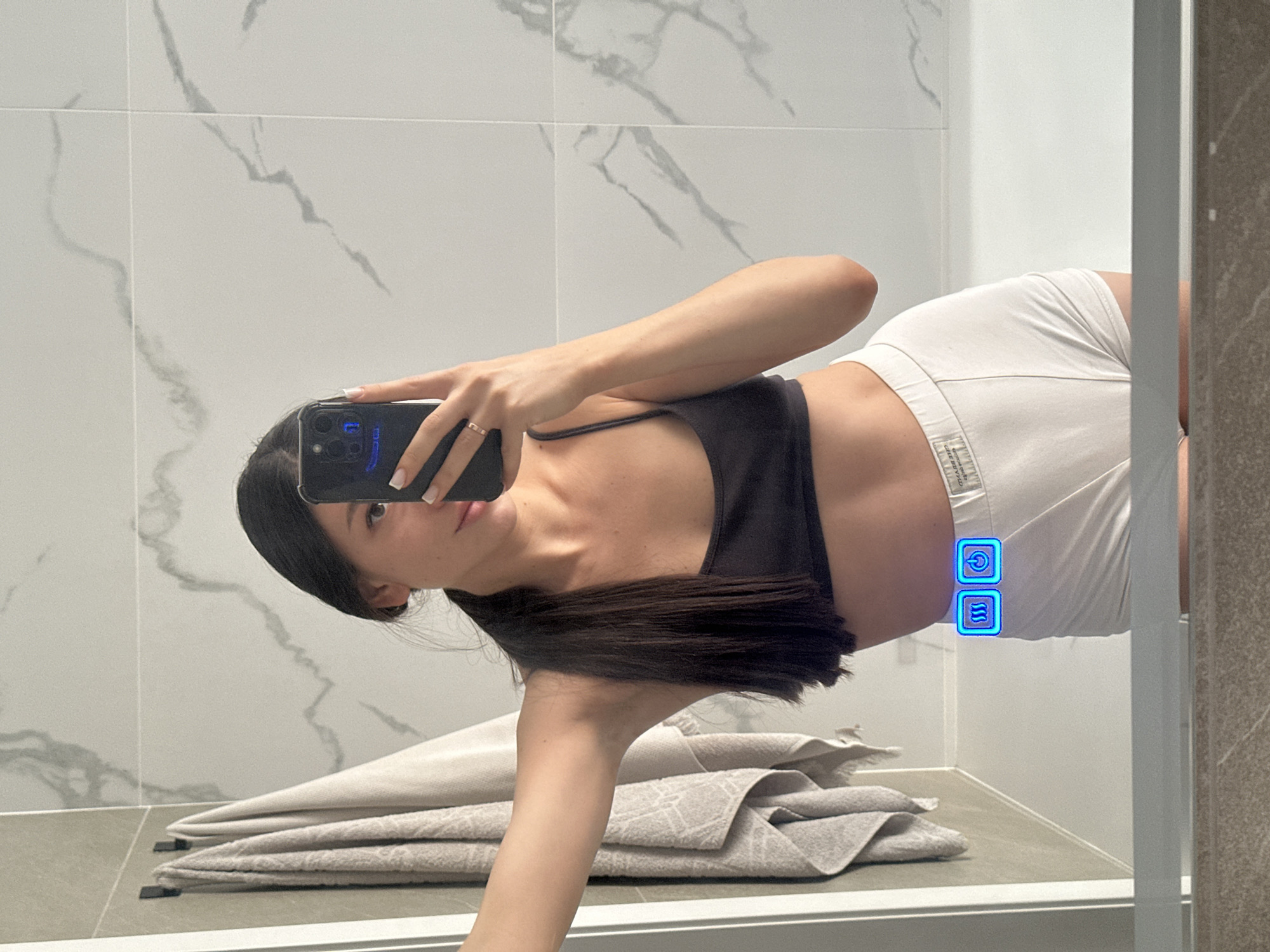 A young woman with dark hair pulled into a ponytail, wearing a black sports bra and white shorts, is taking a mirror selfie in a bathroom. She is holding a smartphone in her right hand, and her left arm is resting on a towel rack. On her shorts, near her hip, there are two illuminated blue icons: one depicting waves of heat and the other a power button. The bathroom has marble-patterned tiles on the walls and a bathtub visible. The image is taken in Эстачи, Russia.
