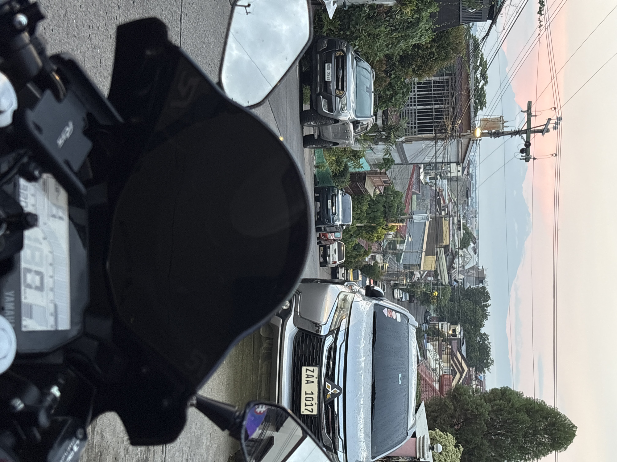 The image captures a view from a motorcycle windshield looking down a street in Davao City, Philippines, towards a distant mountain range under a pastel sky, possibly at dawn or dusk. In the foreground, the black windshield and a portion of the motorcycle's dashboard, displaying digital readouts and the "YAMAHA" logo, are visible.
The street is lined with houses and buildings, many with tiled roofs, indicative of a residential or mixed-use area. Several vehicles are parked or in motion on the road. Closest to the motorcycle are a silver SUV with license plate ZAA 1017 and a grey pickup truck with license plate NDD 2548. Further down the street, other vehicles, including a white pickup truck and what appears to be a jeepney or bus, are visible.
Utility poles with a dense network of wires crisscross overhead, a common sight in urban Filipino landscapes. Lush green trees and dense foliage are scattered throughout the scene, softening the urban environment. The distant mountain range, with soft, undulating peaks, provides a natural backdrop to the city's sprawl. The overall impression is a typical street scene in a Philippine city, with a blend of urban infrastructure, residential housing, and natural scenery. The presence of specific license plate formats (NDD and ZAA) can potentially help pinpoint a more specific location within Davao City, as these formats are used in the Philippines.