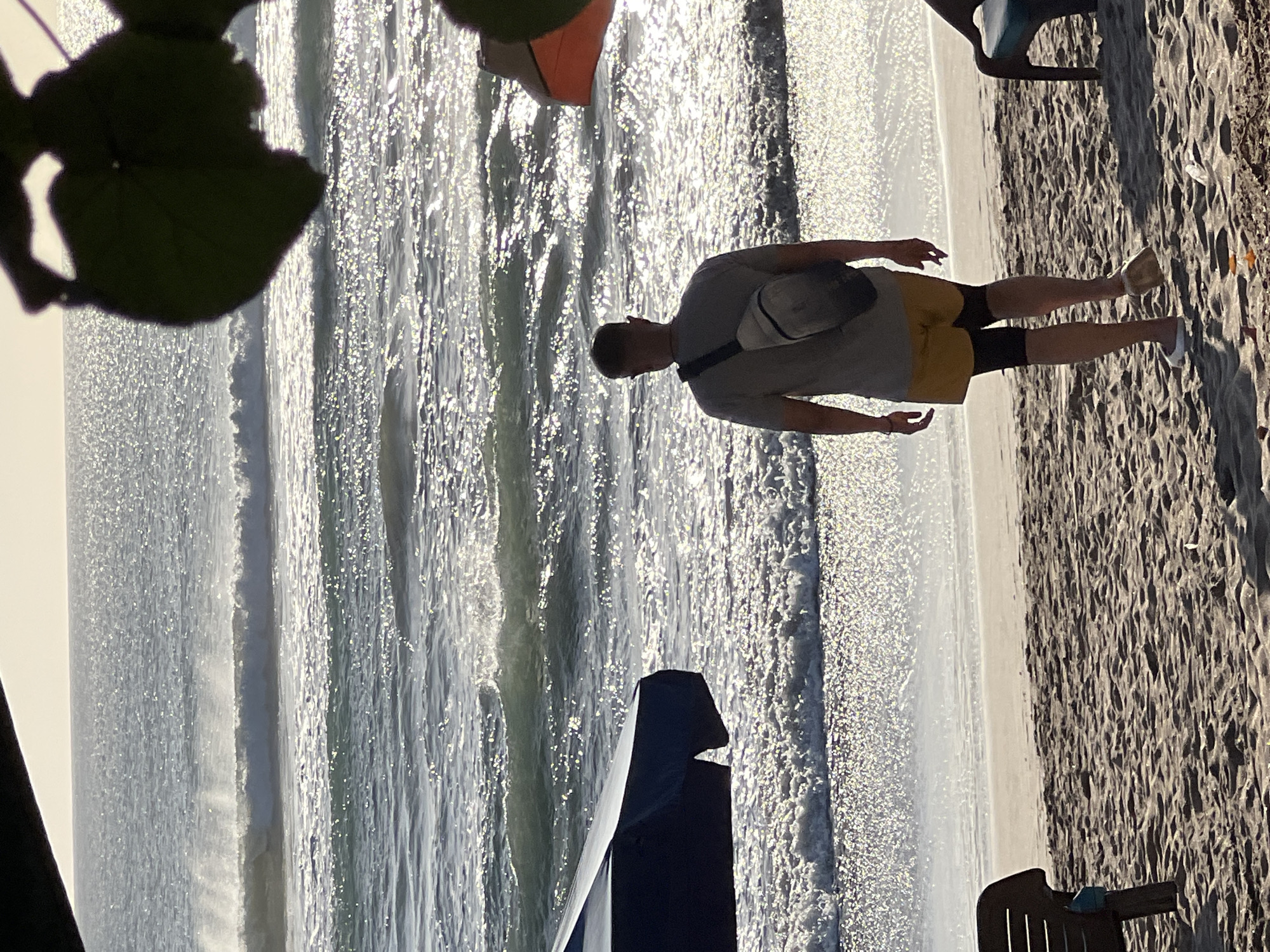 A man walks on a sandy beach in Kuta, Indonesia, facing away from the camera towards the ocean. He is wearing a grey t-shirt, yellow shorts, and black compression leg sleeves, with white slip-on shoes. A light-colored sling bag is worn across his chest. The ocean has moderate waves and reflects the sunlight, creating a glittering effect on the water's surface. To the left, a dark blue beach umbrella is partially visible, and to the far right, a section of a beach chair can be seen. The sky is bright and hazy, suggesting a sunny day. In the foreground, sand is textured with footprints and scattered debris.